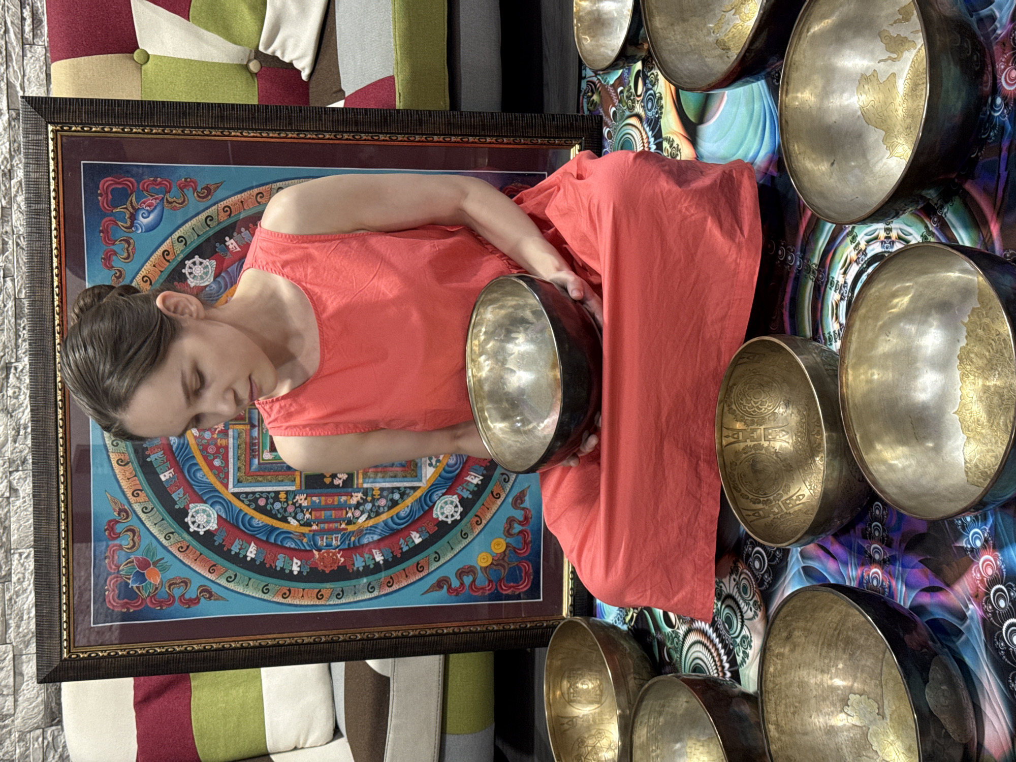 A woman in a reddish-orange dress is seen in what appears to be an indoor setting. She is holding a large metallic bowl in front of her. To her left is a framed piece of artwork with a complex, colorful, circular design. Around the woman and the artwork are several singing bowls of various sizes, set against a multi-colored, psychedelic-patterned background. The image is taken from a high-angle perspective.
While the city of Tashkent, Uzbekistan is mentioned in the prompt, there is no information available to confirm or support the location being in Tashkent.