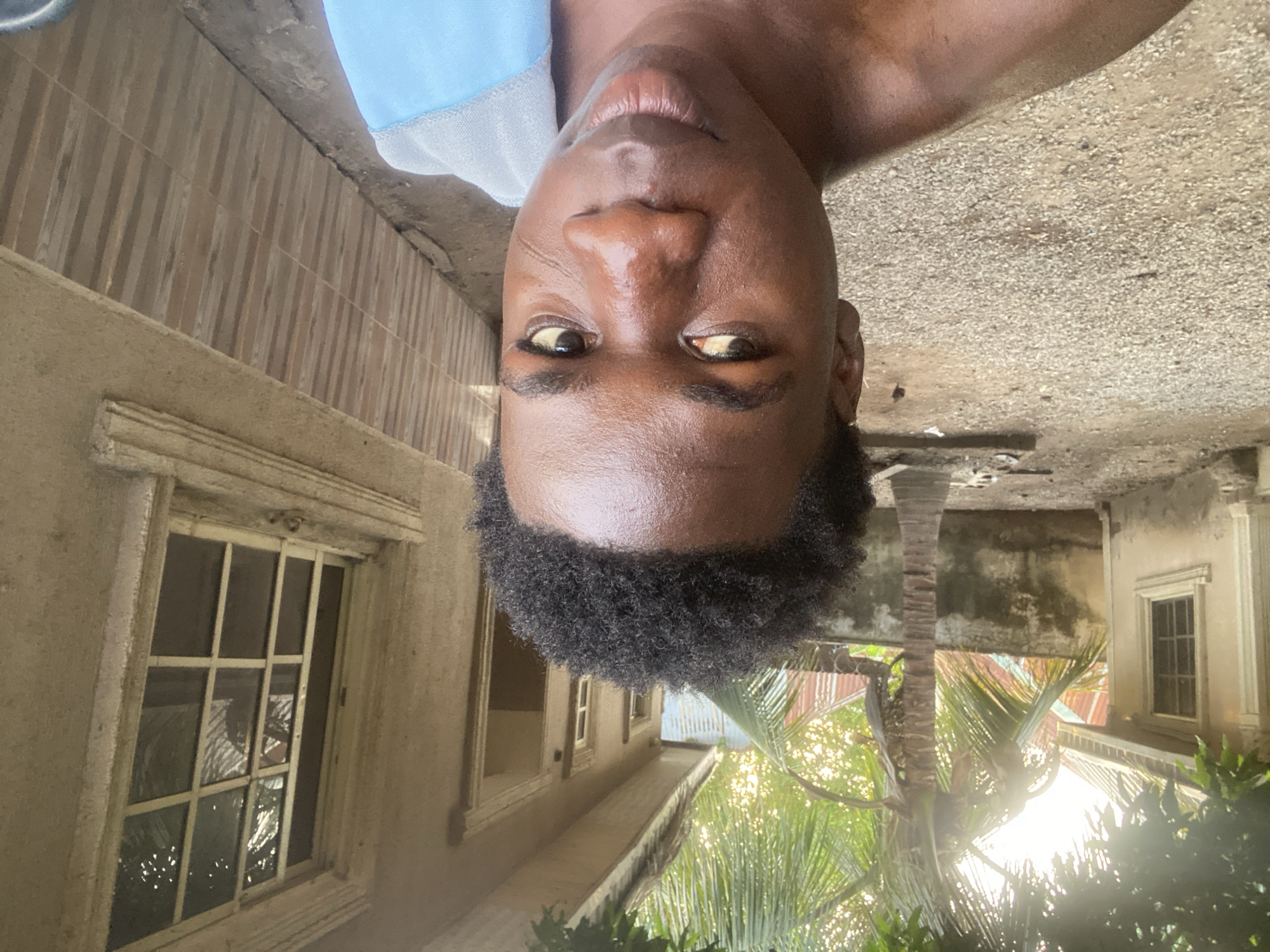 An upside-down, ground-level, medium shot reveals a man’s face. His dark, curly hair tops his forehead. His skin appears oily, reflecting light. He’s wearing a blue/gray top. Behind him, partially obscured by palm tree fronds, is a building with old windows and peeling paint. The ground, likely concrete, is visible in the upper portion of the frame. This image appears to be taken in Mararaba, Nigeria, based on location context.