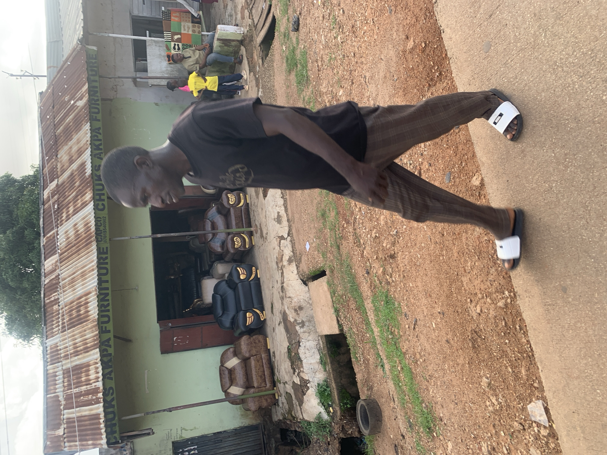 The image captures a street scene in Jos, Nigeria. The primary subject is a man wearing a dark t-shirt and checkered pants, walking on a paved path next to a dirt road. He is seen from the waist down, with his right foot shod in a white slide sandal.
Behind the man, to his left, is a building that appears to be a furniture shop. The shop has a corrugated metal roof and a green facade with signage. Visible text on the building reads "CHUKS AKPA FURNITURE" vertically. In front of the shop, a variety of furniture is displayed, including what looks like leather sofas and chairs in brown and black. Some of these furniture pieces are stacked or placed outdoors.
Further in the background, other people are visible, including individuals in yellow and light-colored clothing, suggesting other activities or patrons associated with the area. The setting appears to be an outdoor commercial space, possibly a market or a street with various businesses. The ground is a mix of dirt, gravel, and some patches of grass, with a visible drainage channel. The lighting suggests it might be daytime, possibly late morning or afternoon, with overcast or diffused light. The overall impression is one of everyday commerce in a Nigerian urban or semi-urban environment.