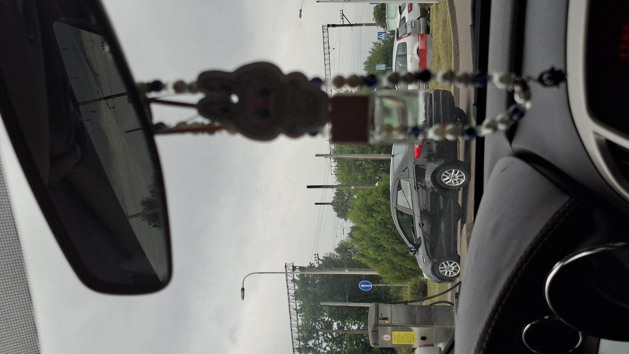 The image, taken from inside a vehicle in Smalyavichy, Belarus, shows a view through the windshield and rearview mirror. In the foreground, the dashboard of the car is visible, with a dark, textured surface. A car air freshener, featuring a cartoonish figure and a small glass bottle, hangs from the rearview mirror. The rearview mirror reflects a cloudy sky and distant trees.
Outside the car, a black sedan is parked. To the left of the sedan is a metal kiosk or machine, possibly a payment station or service point, with yellow and white signage. Further in the background, there are trees, utility poles, and overhead power lines, indicative of an urban or semi-urban environment. A blue circular sign with a white arrow is visible on a pole. Several other vehicles, including white SUVs or station wagons, are parked or driving in the distance. The overall atmosphere suggests an overcast day.