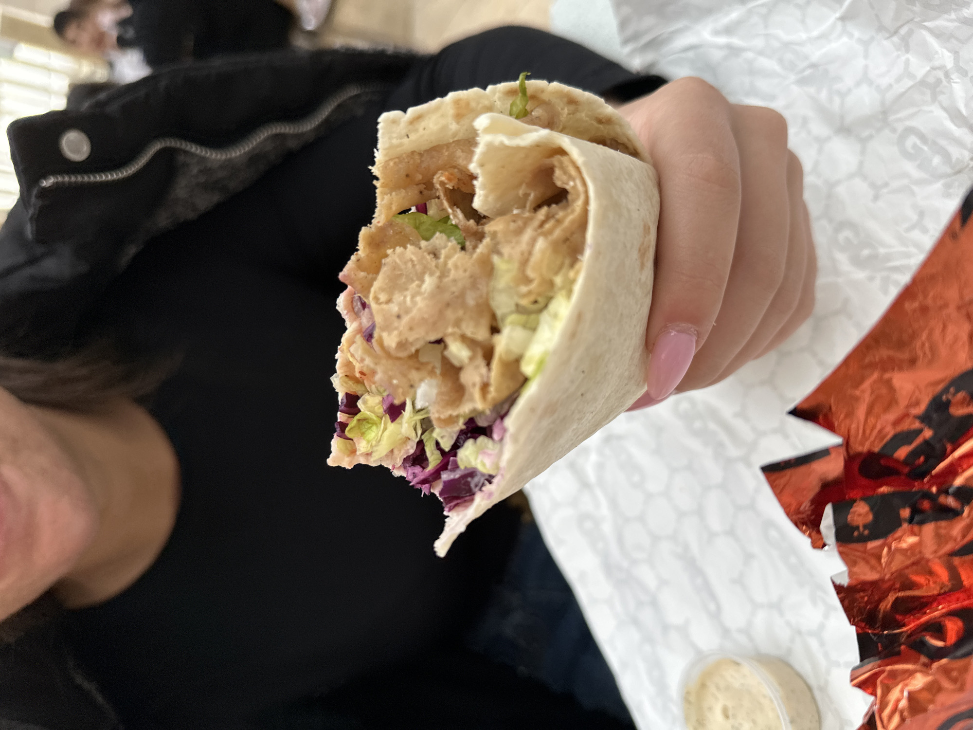 A person is holding a partially eaten wrap. The wrap contains shredded lettuce, red cabbage, and meat. A small container of sauce is visible in the lower right corner. The person is wearing a black top and a black jacket with a zipper. The background includes crumpled white paper and a partially visible orange foil wrapper. The location is unknown and cannot be determined from the image.