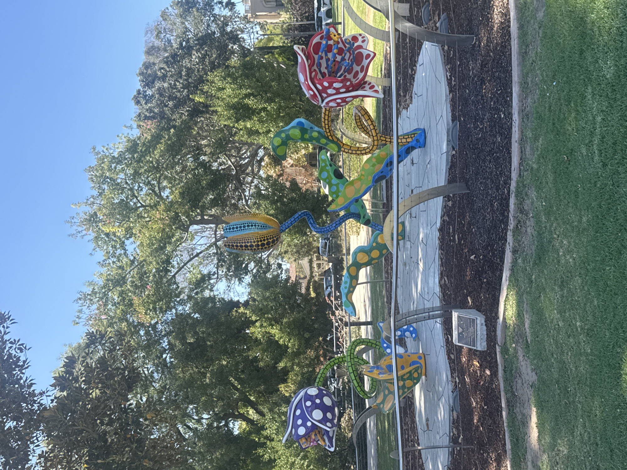 A vibrant, abstract sculpture of flowers with polka dots is displayed outdoors in Beverly Hills, California. The sculpture features large, stylized flowers in red, purple, yellow, blue, and green. The flowers are supported by curving stems and are adorned with numerous dots in contrasting colors. The sculpture is situated on a paved circular base surrounded by dark mulch and a low metal railing. Beyond the sculpture, trees and buildings are visible, suggesting an urban park or public space setting. The sky is clear and blue.