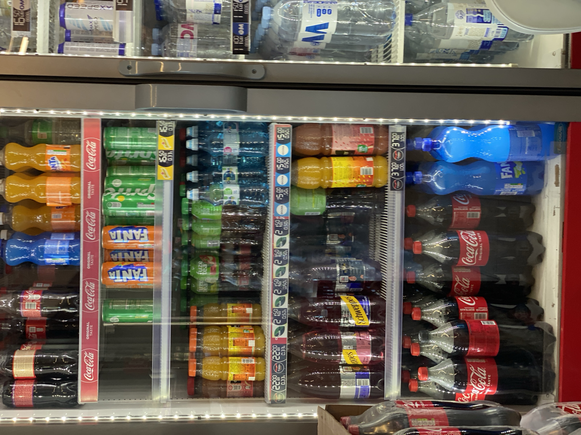 The image shows the interior of a refrigerated display case, likely in a convenience store or supermarket, filled with various beverages. The display case has multiple shelves. The top shelf contains large plastic bottles of Coca-Cola. The second shelf holds cans of Fanta and Sprite, and some other unidentified soda cans. The third shelf features plastic bottles of what appears to be juice or flavored water. The fourth shelf displays bottles of Schweppes, with labels indicating flavors like "Pink Tonic" and "Taste of Blueberry Marula." Below that, a shelf contains large plastic bottles of Coca-Cola. The right side of the display case, visible through a glass door, shows rows of bottled water and other drinks. The lighting appears to be artificial, emanating from within the display case, highlighting the products. The overall environment suggests a retail setting where drinks are kept chilled for sale. There are no people visible in the image. Based on the currency visible on some price tags (LEI, Romanian Leu, which is also the currency of Moldova), and the specific product branding, this location is likely in Chișinău, Moldova.