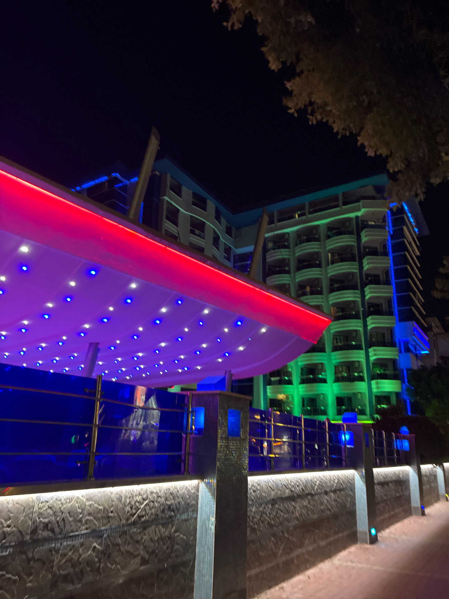 A night exterior shot shows a modern building illuminated with vibrant green and blue lights, located in Alanya, Turkey. A covered outdoor area in the foreground is lit with a red neon border and numerous small blue and white lights on its underside, creating a festive atmosphere. A stone wall with integrated lighting runs along the lower edge of the frame, separating a walkway from the building's elevated area. The building itself is multi-story with balconies and is aglow with a green light illuminating its facade, while vertical blue lights accent its corners. A tree with dark foliage is visible against the night sky in the upper right portion of the image. The scene suggests a hotel or entertainment venue in Alanya, Turkey.