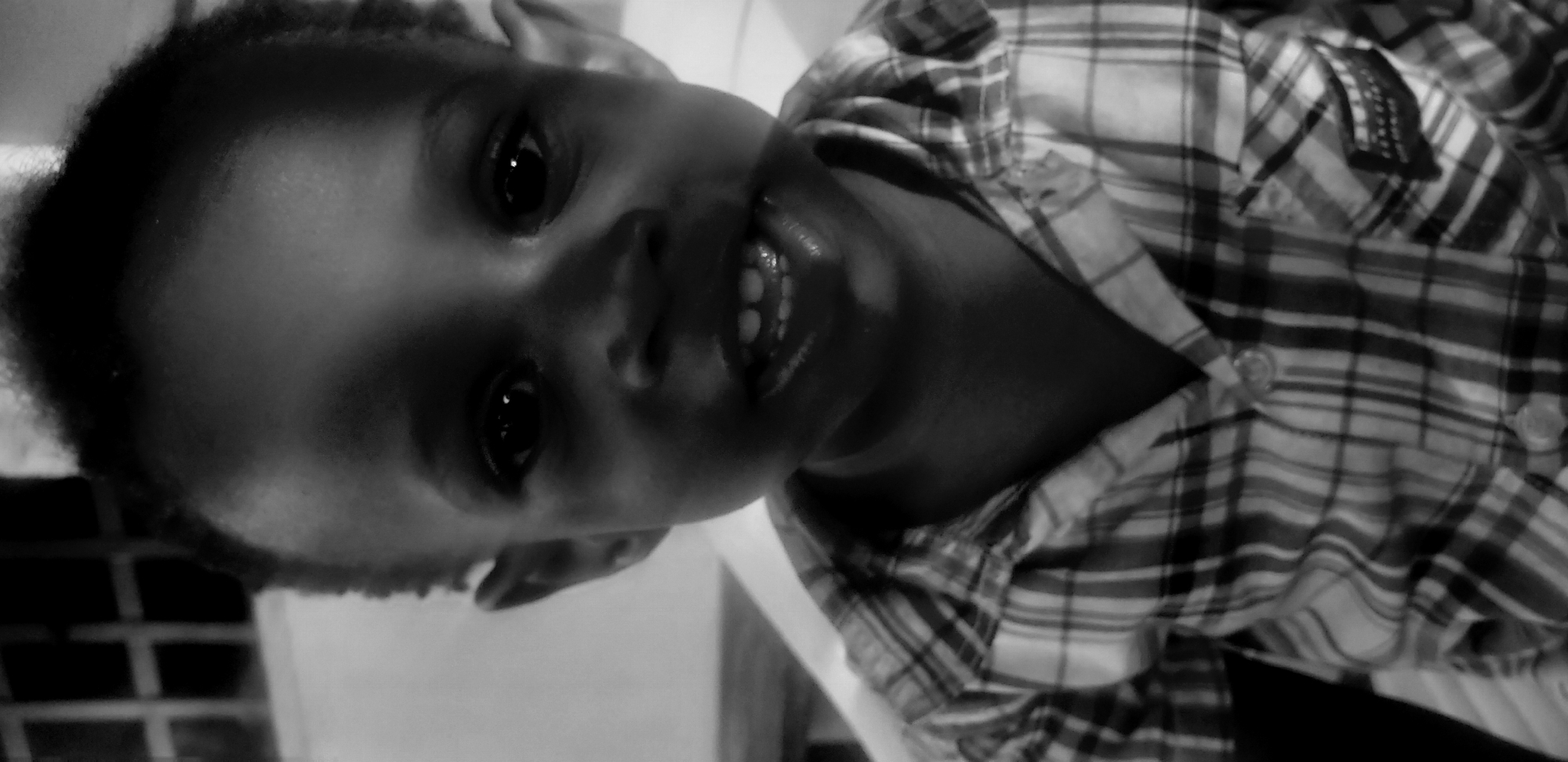 A black and white, medium close-up shot shows a young boy, appearing to be around 3-5 years old, looking directly at the camera. He has dark skin, short, dark hair, and large, dark eyes. His mouth is slightly open, revealing a few small, white teeth. He is wearing a plaid collared shirt with two visible buttons. The background is out of focus, but suggests an indoor setting with some ambient light. The image appears to have been taken in Narayi, Nigeria.