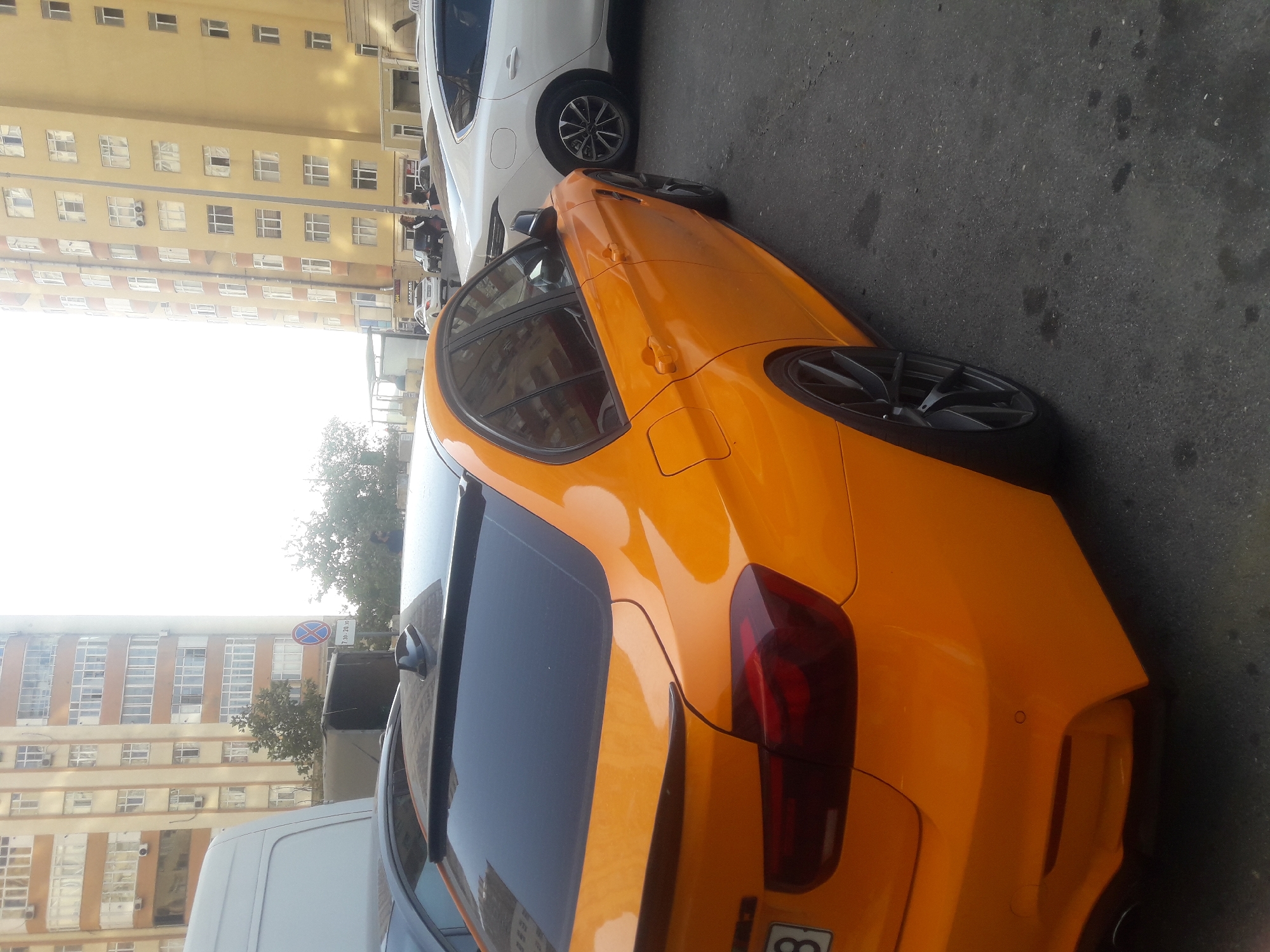 An orange BMW M3 sedan is parked on a street in Baku, Azerbaijan, with the rear passenger side facing the camera. The car has black tinted windows, a spoiler, and dark grey alloy wheels. In the background, there are two tall apartment buildings, one tan and one yellow, with many windows and balconies. A white van is partially visible on the left side of the image, and a white sedan is parked behind the BMW. A street sign with a red circle and a blue cross is visible in the background, along with some trees and foliage. The sky is bright and overcast.