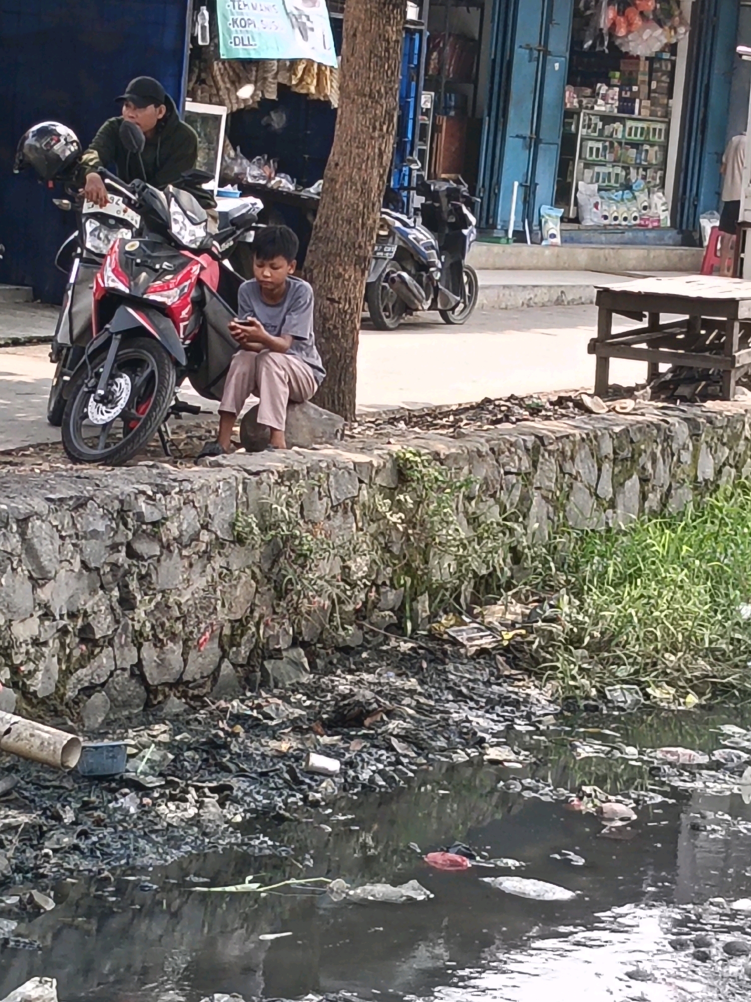 In Belimbing, Indonesia, a young boy sits on a rock by a polluted canal, engrossed in his mobile phone. He is wearing a gray t-shirt and light-colored pants. Next to him, a motorcycle is parked. In the background, a man wearing a black cap and a dark hoodie is leaning against the motorcycle. He is looking to his left. Adjacent to the canal, a stone wall separates the walkway from the polluted water. Behind the wall, a street with shops is visible, with one shop displaying various goods on shelves. The canal itself is filled with dark, murky water and a significant amount of trash, including plastic bottles and bags. Lush green grass grows along the edge of the canal, contrasting with the surrounding debris. A wooden structure, possibly a bench or table, sits on the walkway. The overall scene depicts an urban environment with signs of pollution and everyday life.