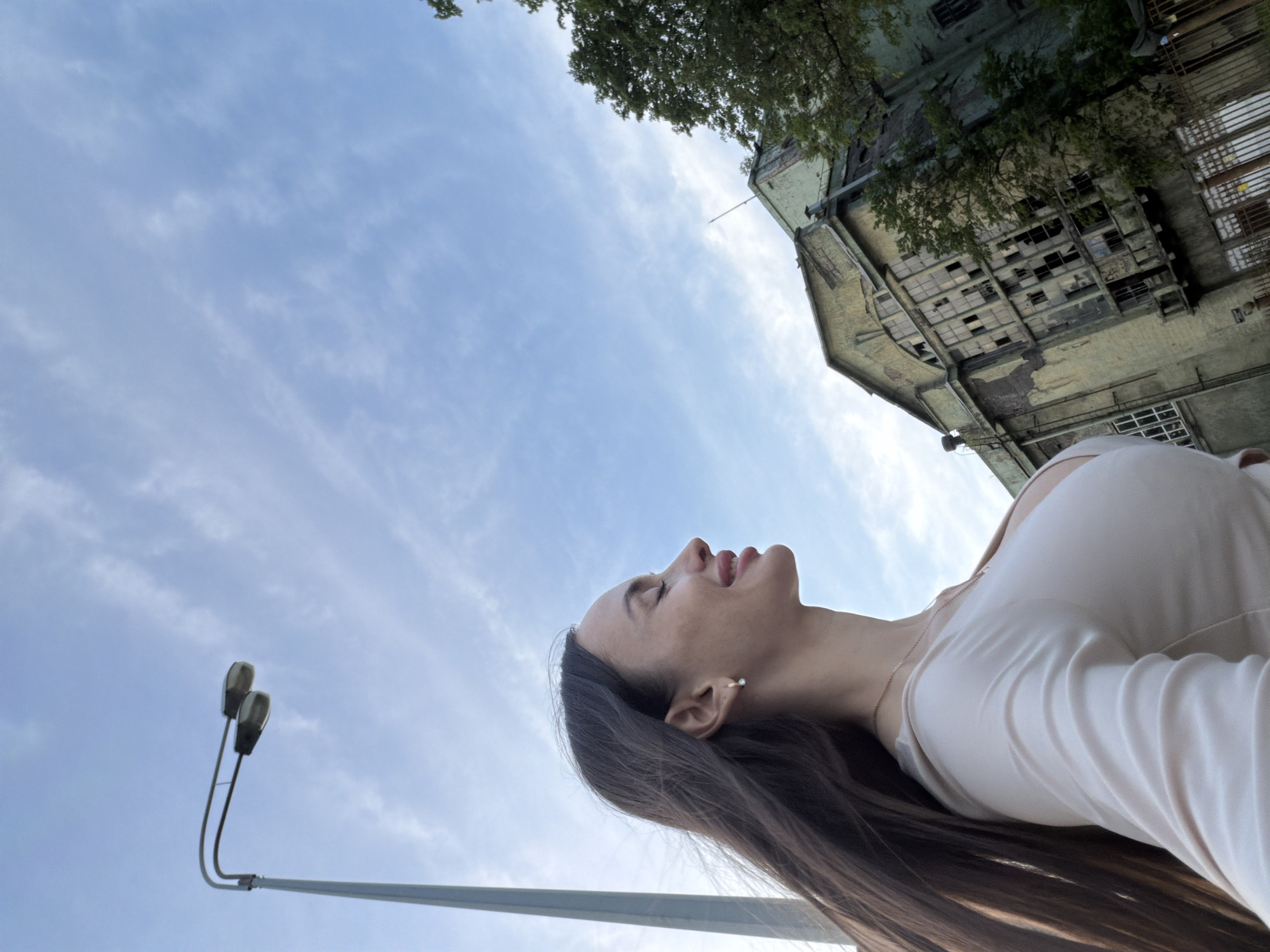 A young woman is depicted in profile, facing right, with her eyes closed and a soft smile, suggesting contentment or enjoyment of the light. She has long, dark brown hair flowing behind her and wears a light-colored, long-sleeved top with a visible neckline and a thin gold chain. A small, white earring is visible in her right ear.
The scene is set outdoors under a light blue sky with scattered, wispy white clouds. A tall, grey metallic lamppost with two attached light fixtures extends from the left side of the frame into the upper left quadrant. In the background, a large, multi-story building with distinct industrial architecture occupies the right side of the image. The building is in a significant state of disrepair, with a faded, peeling greenish-yellow facade, much of its plaster missing, exposing underlying structures. Numerous windows are broken, boarded up, or entirely absent, particularly in a large, grid-like section towards the center-right of the building. A prominent gable structure is visible at the top of the building. Green foliage from trees partially obscures the building's upper right section. A dark metal fence or gate is visible at the bottom right, in front of the dilapidated building.
The building is identifiable as part of the former "Red Triangle" (Красный Треугольник) factory complex, located along the Obvodny Canal Embankment in Saint Petersburg, Russia.