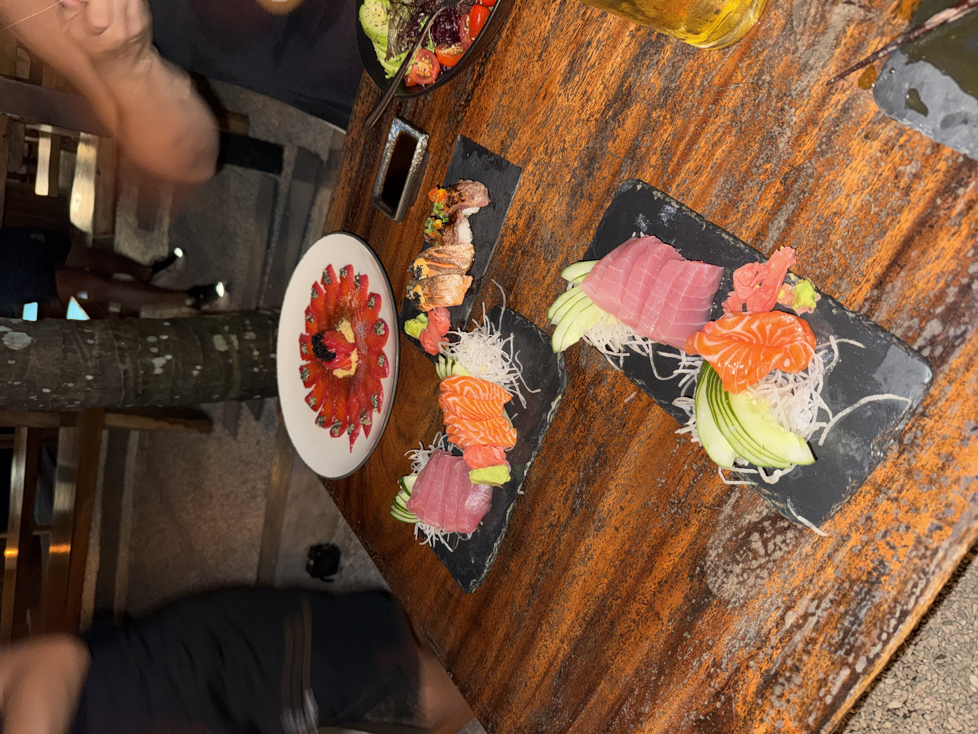 Captured from an overhead perspective, the image shows a diverse culinary spread on a weathered wooden table, possibly in an outdoor dining setting. Visible are plates of sushi and a salad, indicative of a meal being shared. The arrangement of the food suggests a casual dining experience, likely in a restaurant or eatery.
Although a precise location cannot be determined, the prevalence of the meal and its style of cuisine implies it may be a restaurant or open-air setting. People appear in the image, however their faces are not clearly visible. The general dining environment suggests that the city of Kuta Selatan, Indonesia can be described as a popular tourist destination with a vibrant culinary scene.