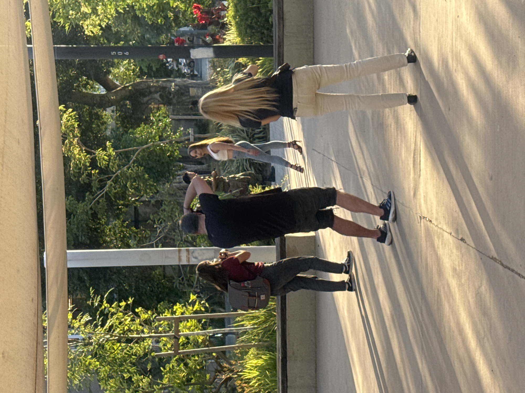 This image captures an outdoor scene in Tel-Aviv, Israel, during what appears to be the golden hour of a sunny late afternoon or early evening, evidenced by the warm, directional light and long shadows stretching across the concrete ground.
The primary activity taking place is a photoshoot or content creation. Five individuals are visible. In the foreground, a person with long dark hair, wearing a maroon t-shirt and dark pants, and carrying a grey backpack, is holding a camera or phone, seemingly capturing the scene. To their right, a man in a black t-shirt and dark shorts is also holding a camera, actively photographing a woman. This woman, with long brown hair, is wearing a white crop top and light blue jeans with high heels, and is posing for the camera. Behind her, another woman with long blonde hair, dressed in a black top and light-colored pants, stands observing or waiting. On the far right, the lower legs of a fifth person in light pants and black shoes are visible.
The setting is an outdoor public space, likely a promenade or paved area bordered by lush green foliage and trees. Red flowers are visible among the greenery in the background. A dark horizontal bar, possibly a fence or railing, runs across the mid-ground, displaying the numbers "506". The overall atmosphere suggests a focused, creative endeavor taking place in a vibrant, sunlit environment.