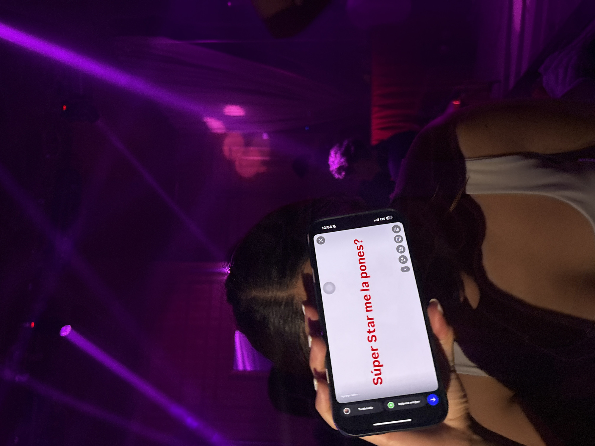 A woman is holding a cell phone in a dimly lit environment with purple lighting, possibly a nightclub. The phone screen displays the words "Super Star me la pones?" in red text. The environment appears to be a crowded setting with indistinct figures in the background. The location is not specified in the picture.