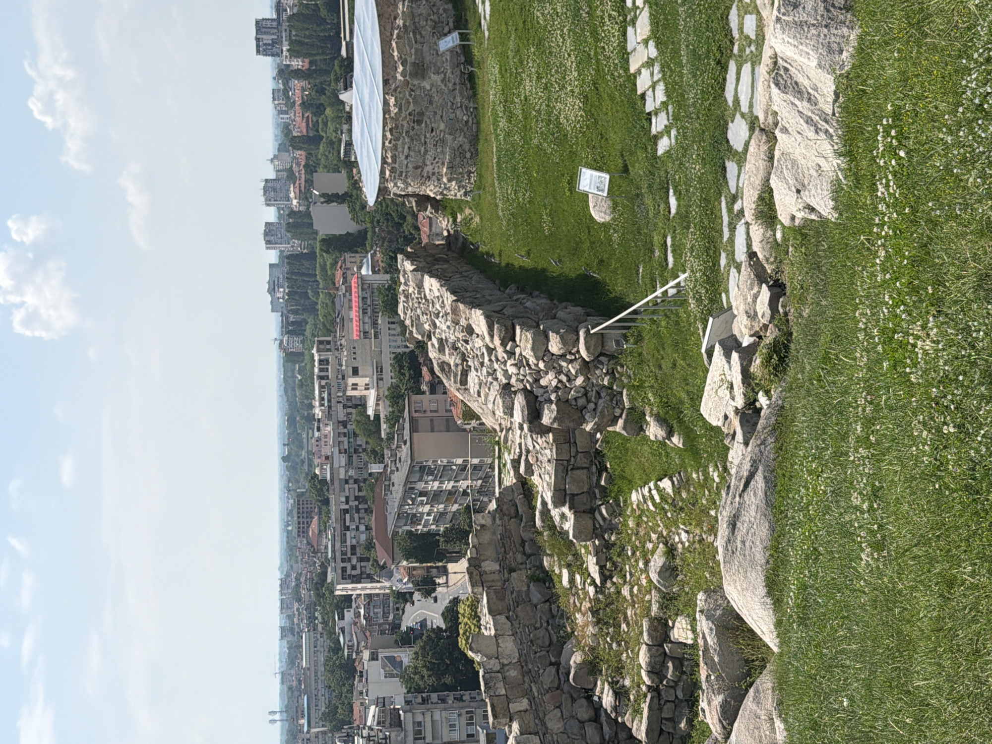This image depicts a panoramic view from an archaeological site on a hill overlooking the city of Plovdiv, Bulgaria, under a bright, partly cloudy sky.
In the foreground, a verdant grassy slope extends across the lower portion of the frame, interspersed with patches of white clover flowers and large, light-colored boulders. Ancient stone ruins, consisting of rough-hewn, irregularly shaped stones, form walls and structures. On the left, a segment of a substantial stone wall runs roughly parallel to the viewer. To the right, another section of a stone wall is visible, and further right, a prominent circular stone structure, possibly a tower or a building foundation, is covered by a modern, translucent, dome-shaped protective roof. A metal handrail is partially visible on the far left, extending downwards. Two white information signs with dark text are positioned on the grass near the ruins. A pathway made of rectangular flagstones is visible on the right, curving away from the circular structure.
The midground features a dense urban landscape of Plovdiv. A network of buildings, ranging from older residential structures with red-tiled roofs to multi-story modern apartment blocks, stretches across the scene. A paved road with a few parked and moving vehicles is visible winding through the buildings in the middle-left. Extensive green tree cover is interspersed throughout the city, notably along streets and between building clusters.
In the background, the city extends towards a hazy horizon. Distant, taller buildings with flat roofs are discernible against the skyline, and communication towers are faintly visible on the far left horizon. The sky is predominantly clear blue with scattered, fluffy white cumulus clouds. No individuals are visible within the frame. The scene is illuminated by bright daylight, suggesting a sunny day.