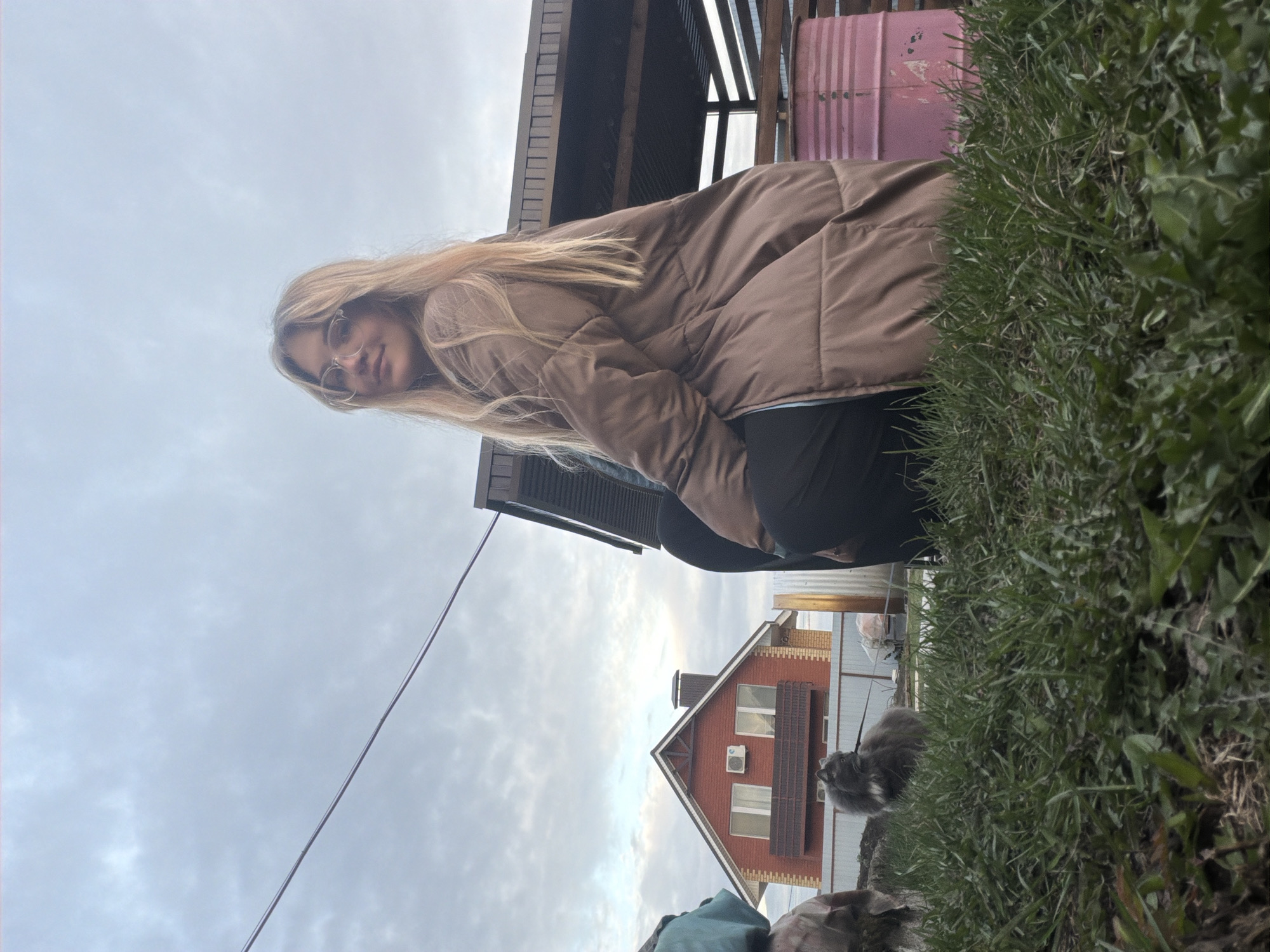 This outdoor photograph, taken in Большая Шильна, Russia, features a young woman and a cat in a residential backyard or garden setting under an overcast sky. The image is captured from a low angle, looking upwards towards the subjects and background elements.
On the right side of the frame, a young woman with long, straight blonde hair and round glasses is visible. She is wearing a brown puffer jacket over dark clothing and is looking directly at the camera with a calm, slightly smiling expression. Her posture suggests she is either bending down or photographed from a significantly lower vantage point.
To the woman's left, a light pink metal barrel is partially visible. Behind her, a rustic wooden structure with dark slats extends across the upper left portion of the image. A thin wire or cable diagonally traverses the grey, cloudy sky.
At the bottom of the image, vibrant green grass and foliage form a natural border. Near the bottom center, a fluffy grey cat with white markings is sitting calmly, likely on a leash, looking towards the left.
In the background, a prominent two-story house with reddish-brown brick walls, white-framed windows, a dark sloped roof, and a chimney is visible. An air conditioning unit is mounted on the side of the house. The weather is cloudy, providing soft, diffused lighting, indicating it is daytime. No discernible text is visible in the image.