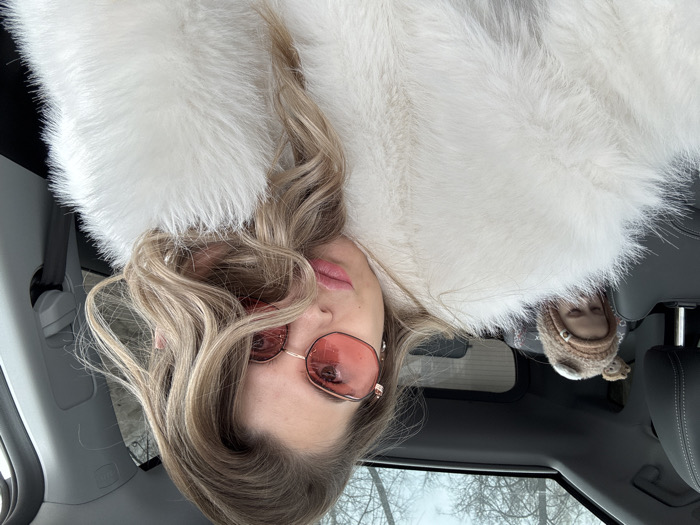 The image depicts two individuals inside a modern vehicle. A female adult is prominently featured in the foreground, occupying what appears to be the front passenger seat. She has light-colored, wavy hair, and is wearing large, round, pink-tinted sunglasses with thin gold frames. Her lips are parted, showing pink lipstick, and she is wearing a voluminous white faux fur coat. Her gaze is directed towards the camera.
In the background, a small child is visible in the rear passenger seat, partially obscured by the foreground subject and car seat. The child is wearing a light brown knitted hat with ear-like appendages and appears to be looking downwards or away. A small, light brown stuffed animal is partially visible behind the child's head.
The vehicle's interior features dark-colored seats and a panoramic sunroof, through which bare tree branches and an overcast sky are visible, suggesting an outdoor, possibly cold, environment. No specific architectural or geographical landmarks are present to definitively identify a precise location within the city of Chaikovsky, Russia.