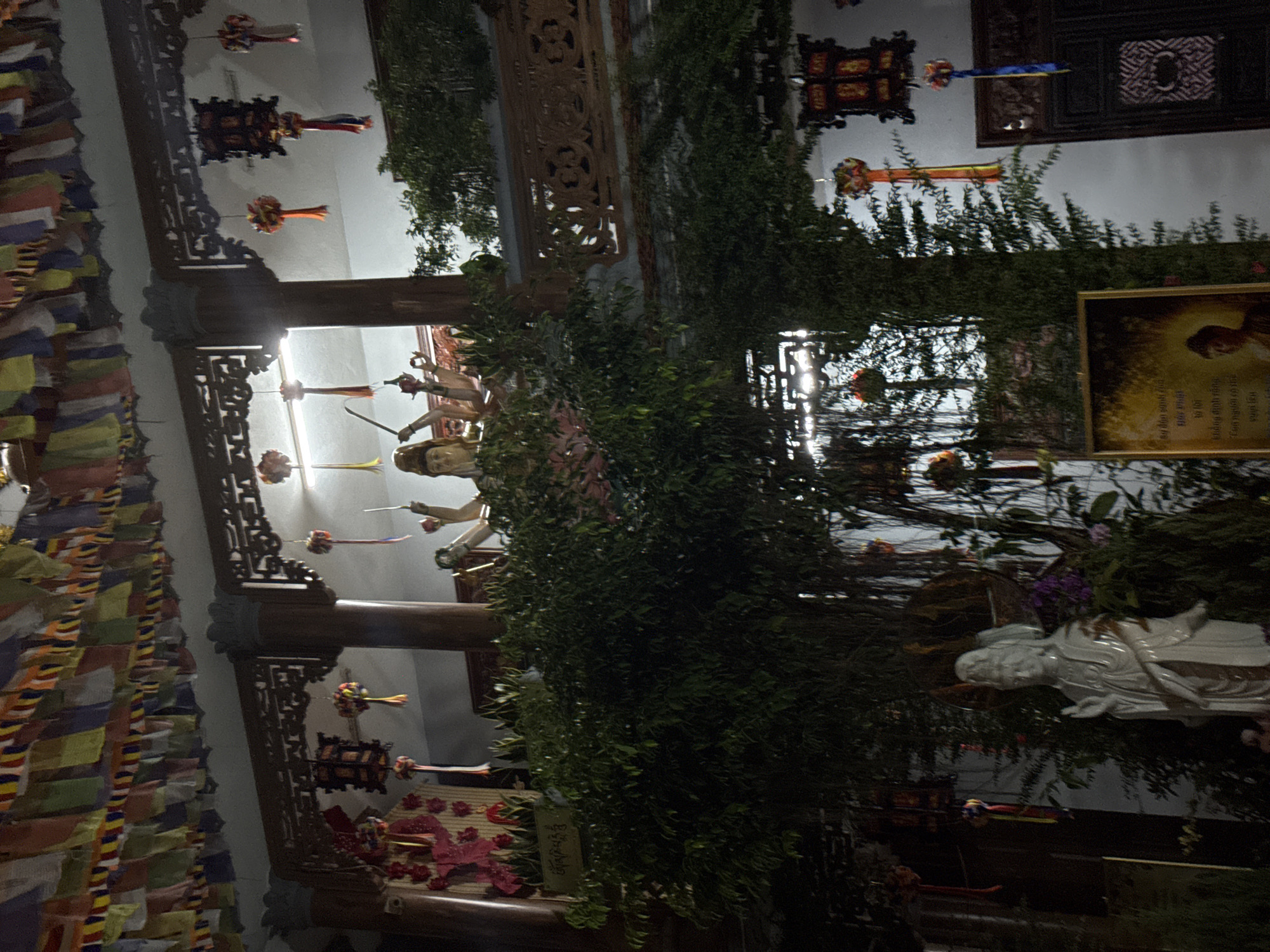 This image shows an interior scene, possibly a temple or religious site, in what appears to be Đà Nẵng, Vietnam. Prayer flags hang on the left, and a series of statues or figures are visible in the background, partly obscured by foliage. Elaborate wooden carvings adorn the architecture. The scene features decorative objects suspended from the ceiling. At the right side, a framed image and a porcelain statue stand among plants.