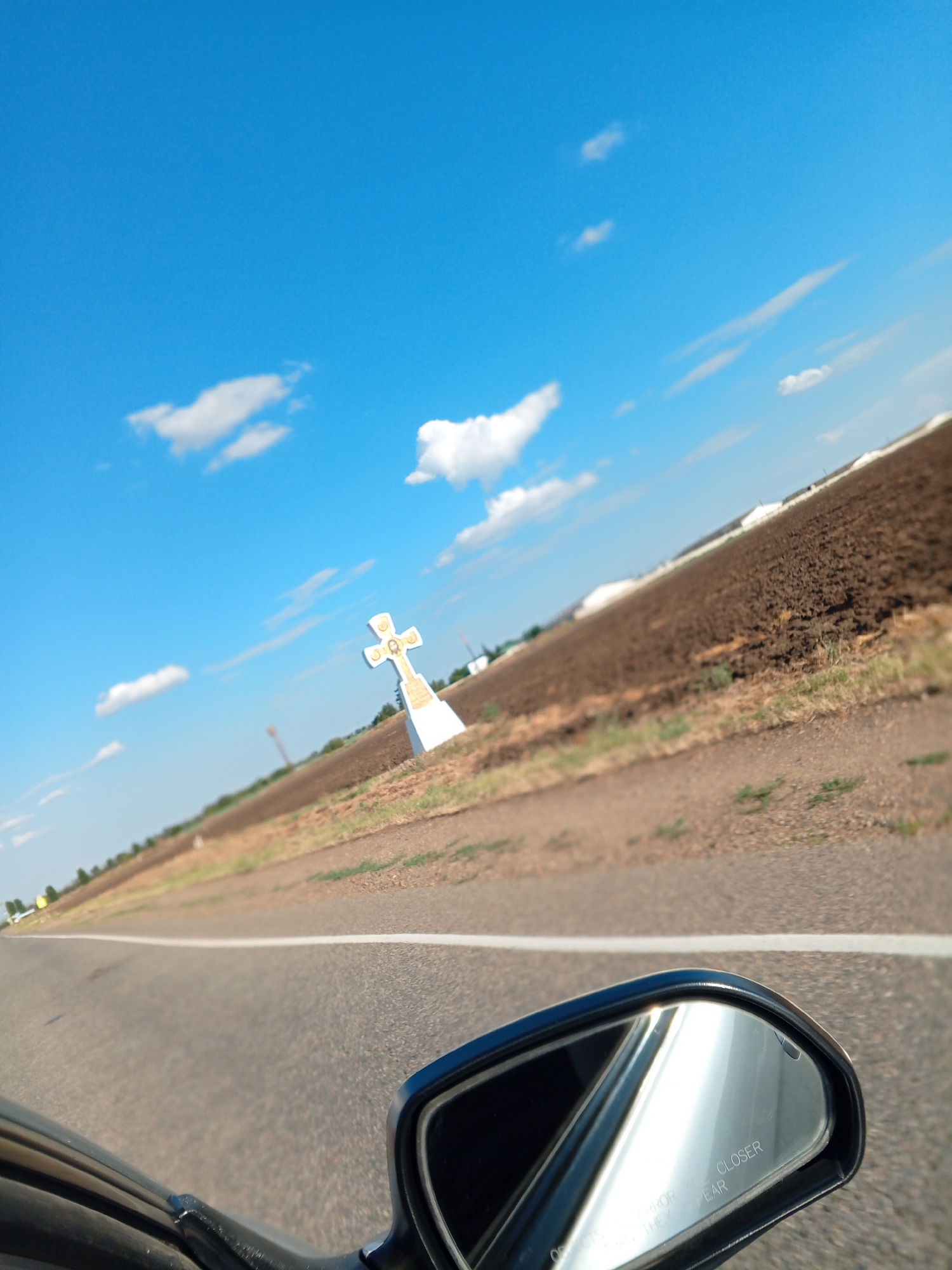 An eye-level, wide-angle shot captures a view from a vehicle, looking out at a rural landscape under a bright blue sky with scattered white clouds. In the middle ground, a prominent white cross with gold accents stands against a backdrop of a plowed brown field. The field is bordered by a dirt road and sparse green vegetation. The road ahead is paved and has a white line marking its center. The lower right corner of the image shows a reflection in the side-view mirror of the vehicle, with the text "Objects in mirror are closer than they appear" visible. The scene appears to be in an open, unpopulated area, possibly along a highway.
