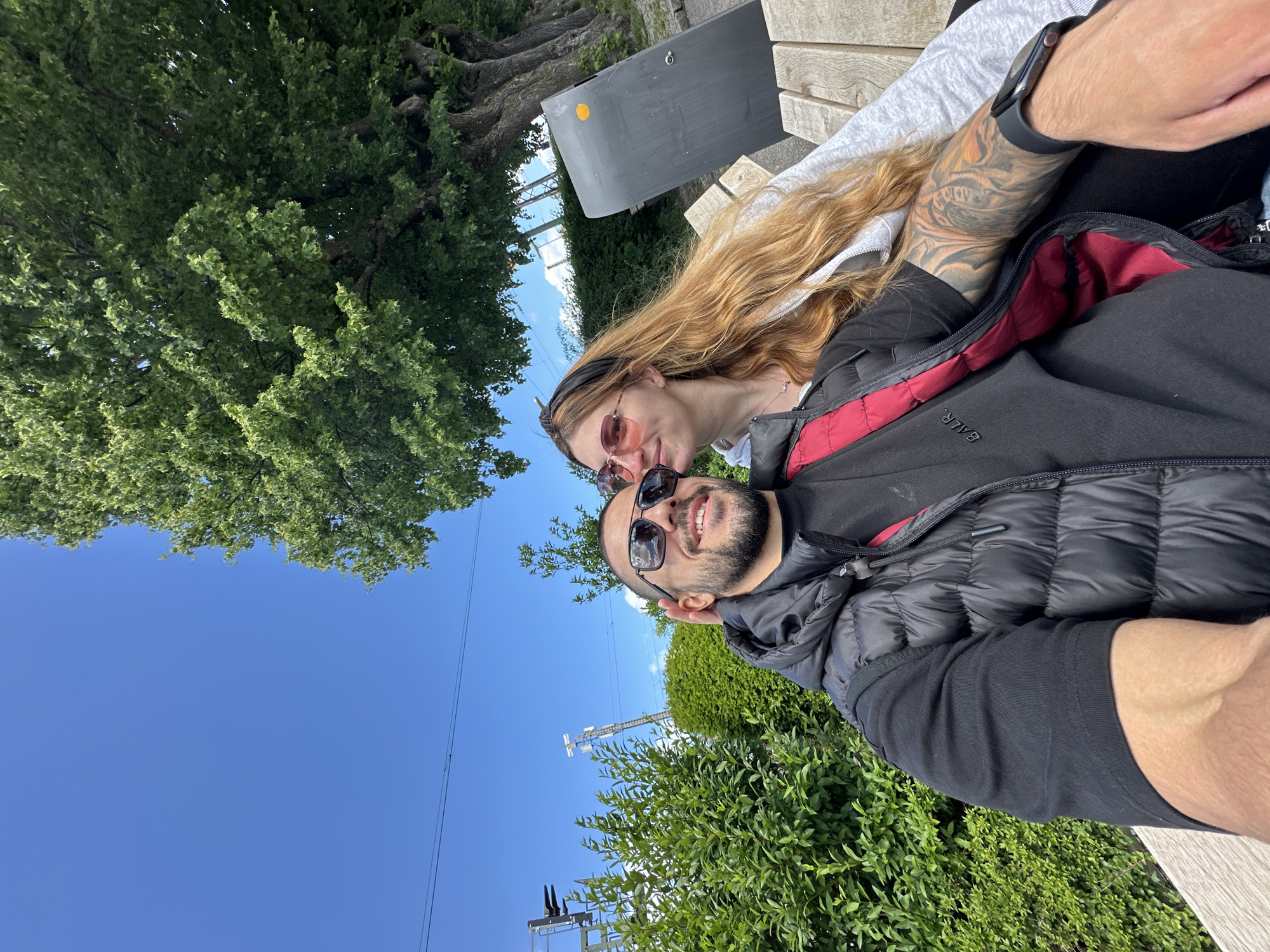 In Starnberg, Germany, a young couple is captured in a selfie on a bright, sunny day. The man, in the foreground, wears sunglasses and a black puffer vest over a black t-shirt. He has a beard and tattoos on his arm. The woman, to his left, also wears sunglasses and has long blonde hair. She is leaning in, resting her head near his. They are seated on a wooden bench, with lush green bushes and trees surrounding them. In the background, a gray trash bin and power lines are visible. The sky is a clear, vibrant blue.