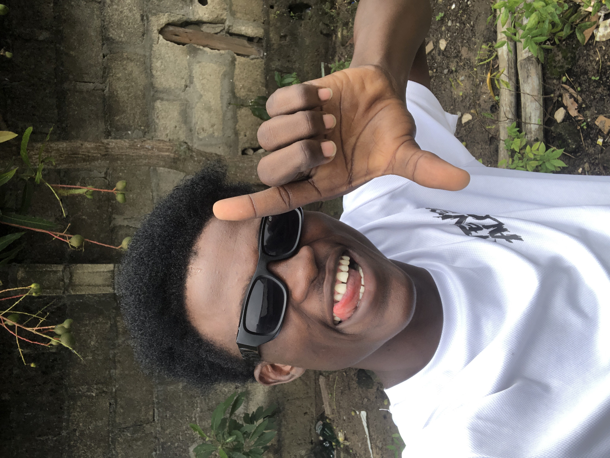 A young Black male with dark, curly hair and black sunglasses is captured in a selfie. He is wearing a white t-shirt with a black graphic on the left side. The man is sticking his tongue out to the left, which is slightly red. His right hand is raised, with his index finger extended upwards and his thumb and middle fingers forming an "L" shape. The background consists of a grey brick wall, a tree with small green fruits, and some green foliage. The ground appears to be dirt with some dry leaves and bamboo-like structures. The scene is outdoors, suggesting a casual setting, possibly in the city of Ado, Nigeria.