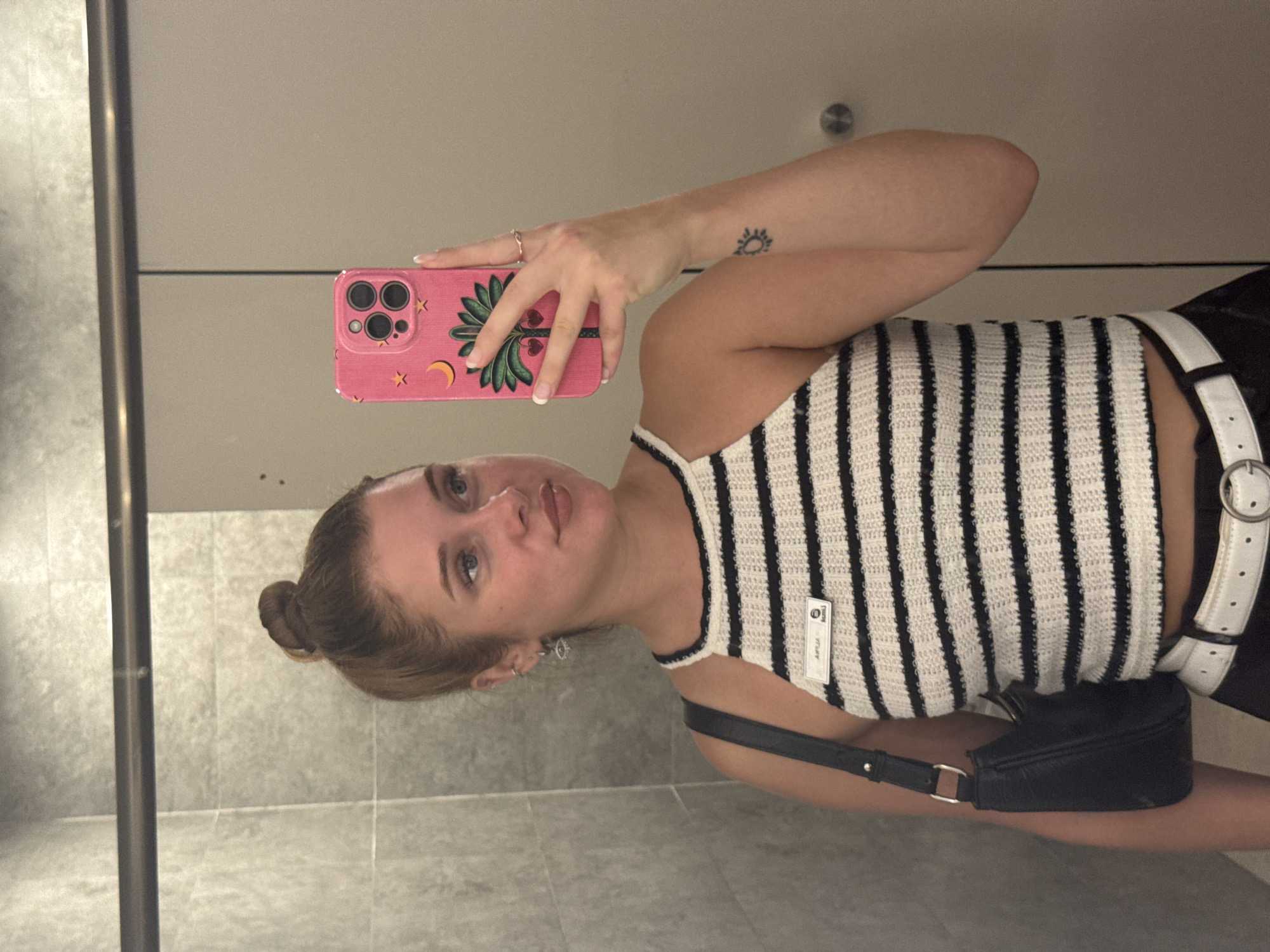 A young woman with her hair in a bun takes a mirror selfie in a well-lit, modern restroom. She is wearing a black and white striped halter top, a white belt, and dark pants, with a black shoulder bag. Her phone, in a bright pink case, is held up to take the photo. The restroom has gray tiled walls and a light-colored door. The woman is wearing earrings and has a small tattoo on her arm. There is a nametag on her top. The scene is set in Aksu, Turkey.