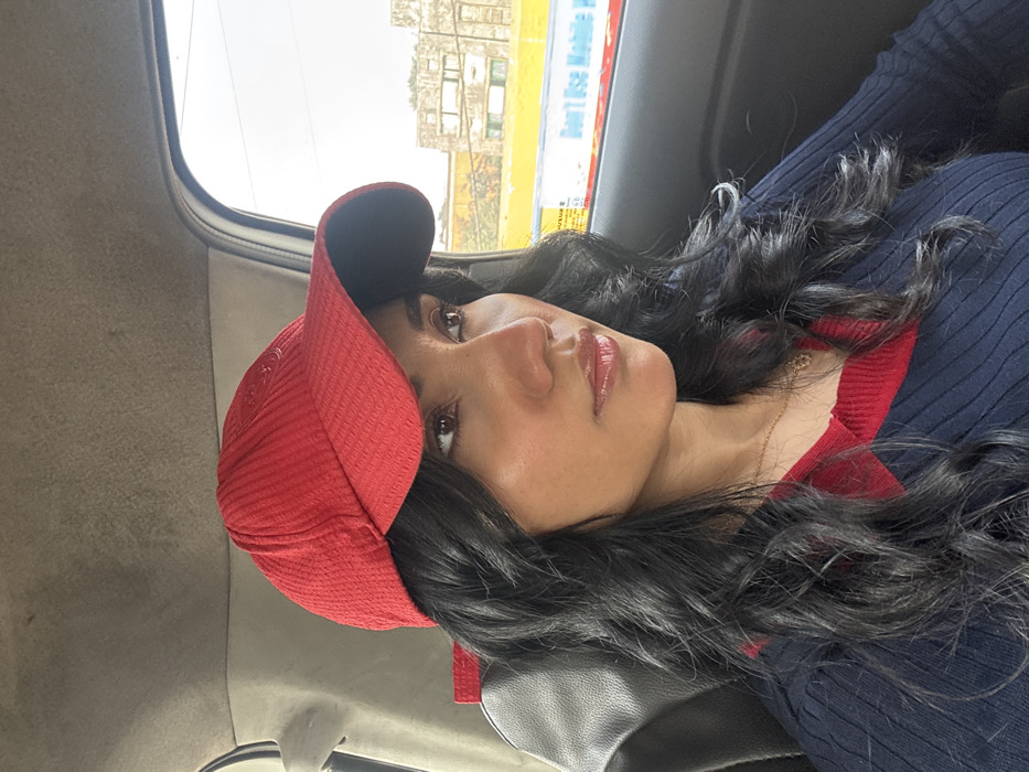A woman wearing a red baseball cap is looking toward the camera while sitting inside a vehicle. She has dark hair, dark eyes, and her lips are glossed. She is wearing a navy blue shirt with red trim at the collar. The background shows a blurry view of buildings and a clear sky through the car window. The interior of the vehicle, including the window frame and seat, is also visible.