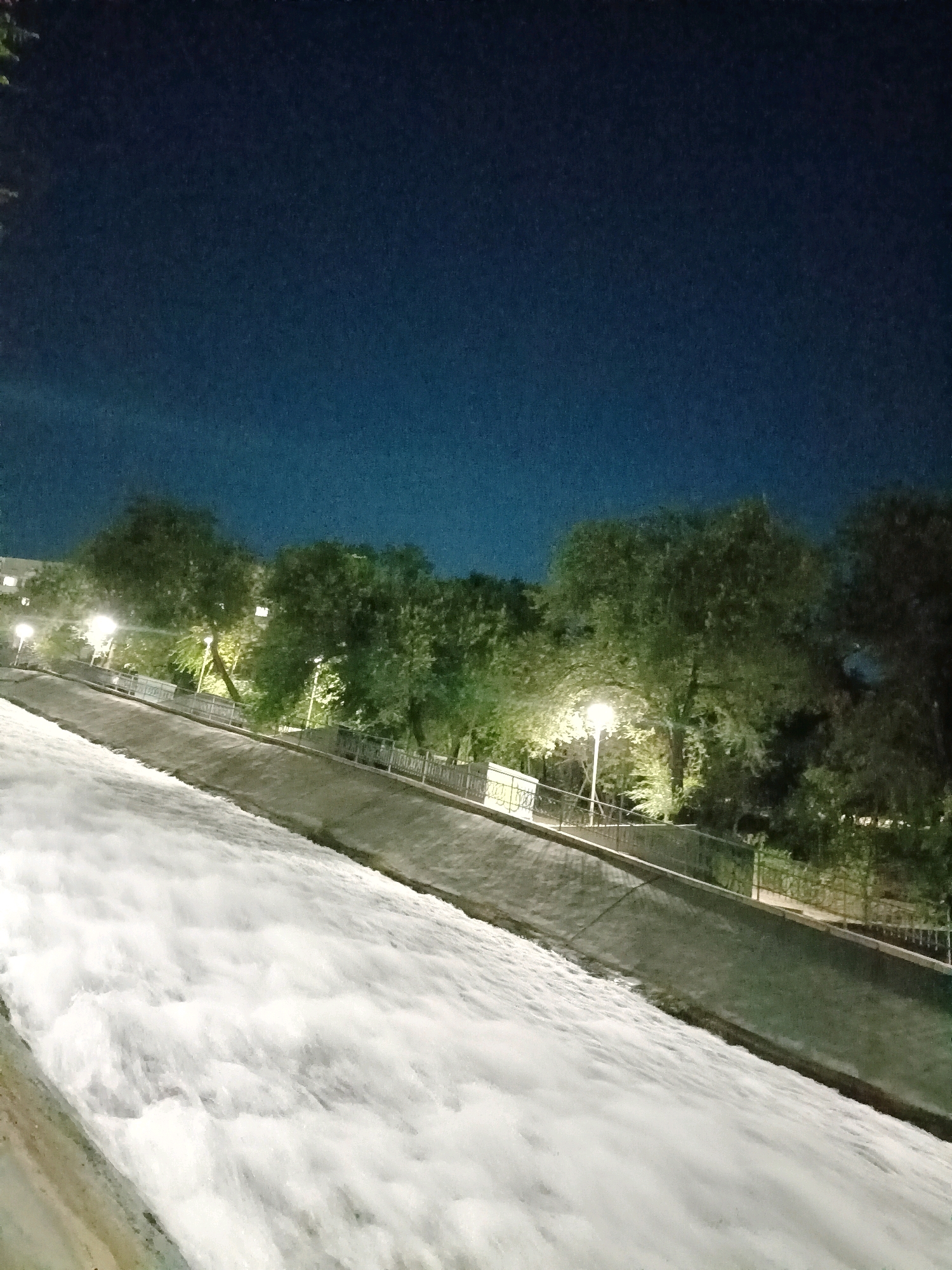 A dimly lit, angled view captures a man-made waterfall or cascade at night. The water flows vigorously down a sloped concrete channel, creating white, frothy waves. Along the upper edge of the channel, a concrete embankment is visible, lined with a metal railing and a fence. Beyond the railing, several trees with lush green foliage are illuminated by streetlights, casting a soft glow. The sky above is a deep, dark blue, transitioning to a lighter shade near the horizon. Buildings with illuminated windows are faintly visible in the background on the left side of the image. The scene appears to be in a park or recreational area, possibly along a river or canal.