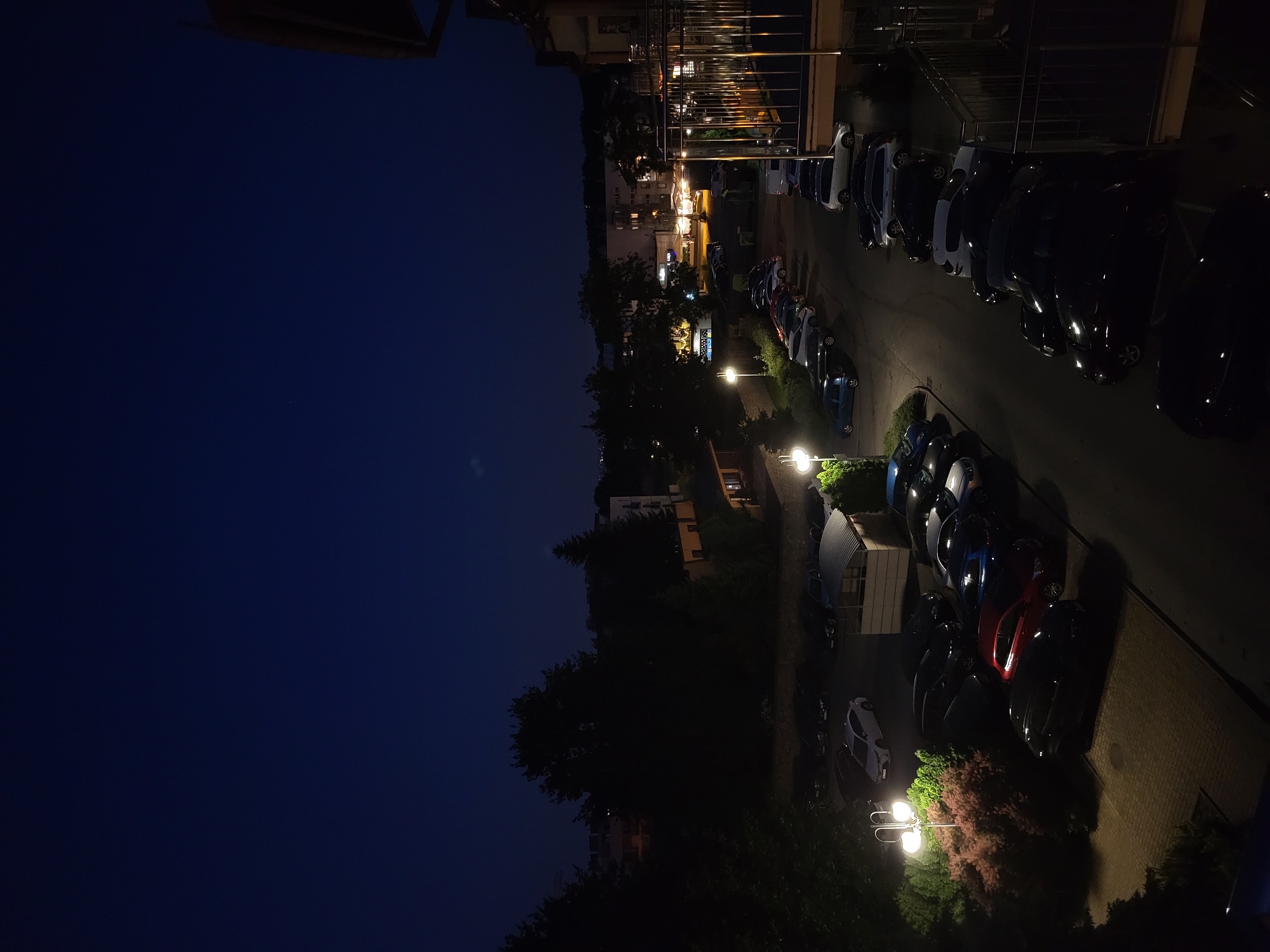 The image is a nighttime aerial view of a parking lot in an urban setting. The sky is dark blue, with a hint of a crescent moon visible. In the foreground, a balcony with metal railings is visible on the right side of the frame. Below, a parking lot is filled with numerous cars of various colors, including white, black, red, blue, and silver. The cars are parked in rows, and some are illuminated by streetlights that cast long shadows.
In the middle ground, trees and bushes are visible, some of which are illuminated by the streetlights. Buildings are visible in the background, some with lights on, suggesting they are occupied. The overall impression is one of a quiet, residential area at night.
The image is clear and sharp, with good detail in the cars and surrounding environment. The lighting is moody and atmospheric, with the streetlights creating a dramatic contrast between light and shadow. The colors are rich and saturated, despite the low light conditions.