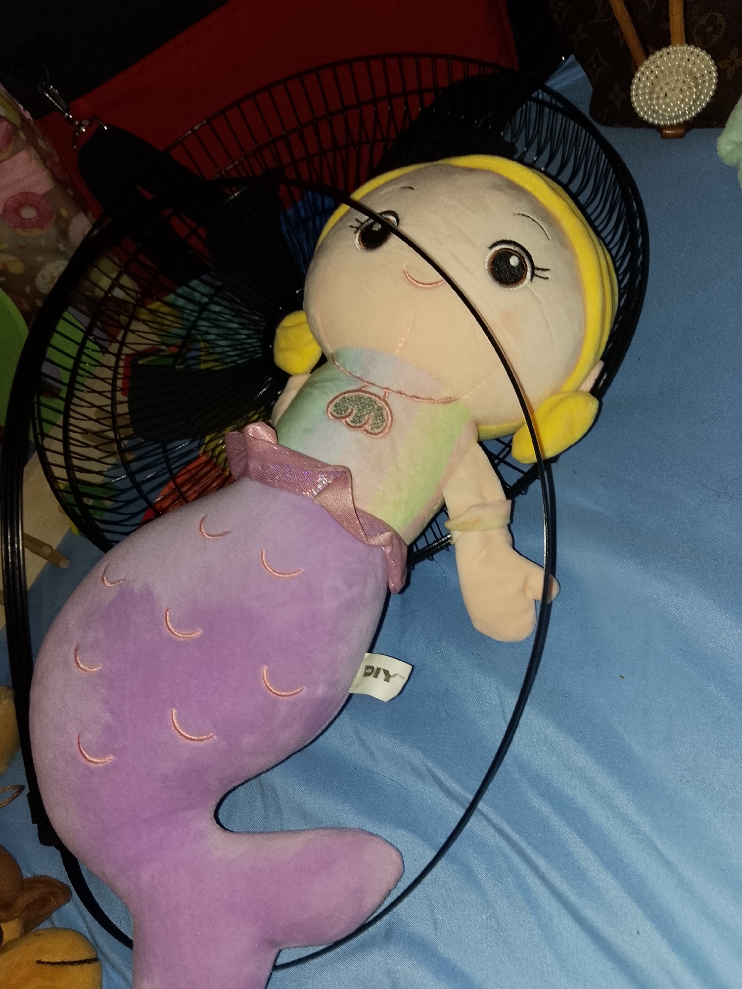 The image captures a plush mermaid doll and an electric fan positioned on a light blue fabric surface, likely a bedsheet, in an indoor setting within Davao City, Philippines.
The central subject is a soft, cuddly mermaid doll. It features a pale yellow or cream-colored head with a friendly expression, large dark eyes, eyelashes, and small eyebrows. The doll has yellow hair, possibly a headband or cap, and light-colored arms. Its upper body is adorned with a pastel, multi-colored top in shades of light green, pink, and yellow, with a small heart or shell design embroidered on the chest. The mermaid's tail is a distinct lavender or purple hue, textured with light pink embroidered crescent shapes that resemble scales. A ruffled, shiny, light purple band separates the doll's top from its tail. A white tag with the black text "DIY" is visible attached to the doll near its tail.
Behind and partially surrounding the mermaid doll is a black electric fan, appearing to be a table or desk model, with its protective grille clearly visible. Through the fan's blades, hints of colorful objects, possibly other toys or fabrics, can be seen in the background.
Additional details in the background include a dark red fabric object in the upper left, and in the upper right, a brown patterned bag, possibly a designer handbag, next to a round, white, pearl-studded decorative item. Another small, brown plush toy is partially visible in the lower-left corner. The scene depicts a static arrangement of objects, with no specific activity or event taking place. The lighting suggests an indoor environment.