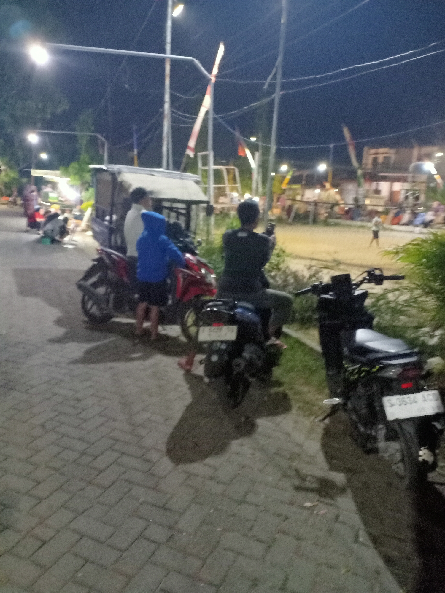 The image depicts a nighttime scene on a paved walkway in Ngraseh, Indonesia, illuminated by streetlights. In the foreground, two motorcycles are parked. The motorcycle on the left is red, and a person in a blue hoodie is standing next to it. The motorcycle on the right is black, with a license plate that reads "S-3634 ACB". A person in a dark shirt is seated on this motorcycle, holding a phone. In the background, a food stall is visible, along with several other people and tents, suggesting a public gathering or market. A flag pole with red and white flags is also present. The overall atmosphere is casual and appears to be a social gathering or a marketplace event after dark.