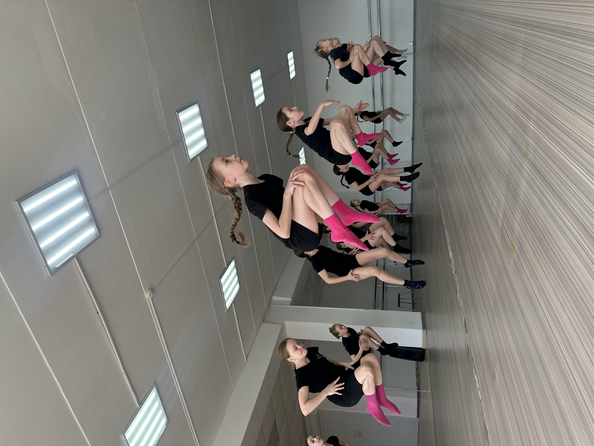 The image shows a group of young girls in a dance studio. The studio has a mirrored wall and bright fluorescent lighting. The girls are wearing black shorts and t-shirts, with some wearing colorful socks. They are seated on the floor in a line, possibly practicing a dance routine. The location is likely Votkinsk, Russia.
