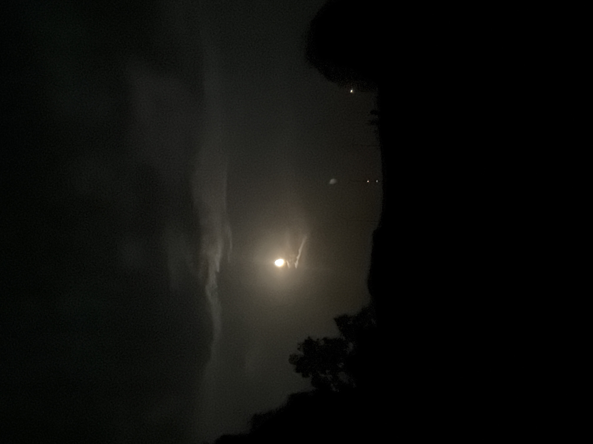 A full moon is visible in a dark, cloudy sky. The moon is partially obscured by thin clouds, creating a hazy glow around it. In the foreground, silhouettes of trees and a low horizon line are visible against the illuminated sky. The scene appears to be an outdoor landscape at night. The location is identified as Creußen, Germany.