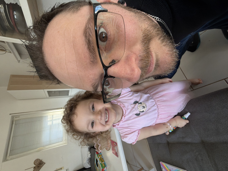The image captures an adult male and a young female child indoors in Trassem, Germany. The adult male, positioned in the foreground, is taking a selfie-style photograph. He is Caucasian, with short brown hair, a light brown full beard and mustache, and wears black-framed, semi-rimless glasses. His brown eyes are open wide and directed at the camera. He wears a dark long-sleeved shirt and a silver chain necklace, with black ear gauges visible.
Standing slightly behind and to the left of the male is a Caucasian female child, estimated to be 3-5 years old. She has curly light brown hair and is smiling broadly, revealing her teeth, while looking directly at the camera. She is dressed in a light pink short-sleeved dress featuring a Minnie Mouse graphic on the chest and a sheer, sparkly overlay on the skirt. She is barefoot. A light pink beaded bracelet adorns her right wrist, and she holds a multi-colored cylindrical marker in her right hand.
The background reveals a residential setting. To the left, a white table holds a light brown decorative coaster and a partially visible patterned box. A gray upholstered surface, likely a couch or cushion, is in the lower-left corner. An angled skylight window with horizontal blinds is visible in the upper-left background, above which hangs a heart-shaped wooden decoration. To the right of the window, light-colored overhead cabinets and a taller white cabinet with a glass panel are present. The floor is light-colored tile or laminate.