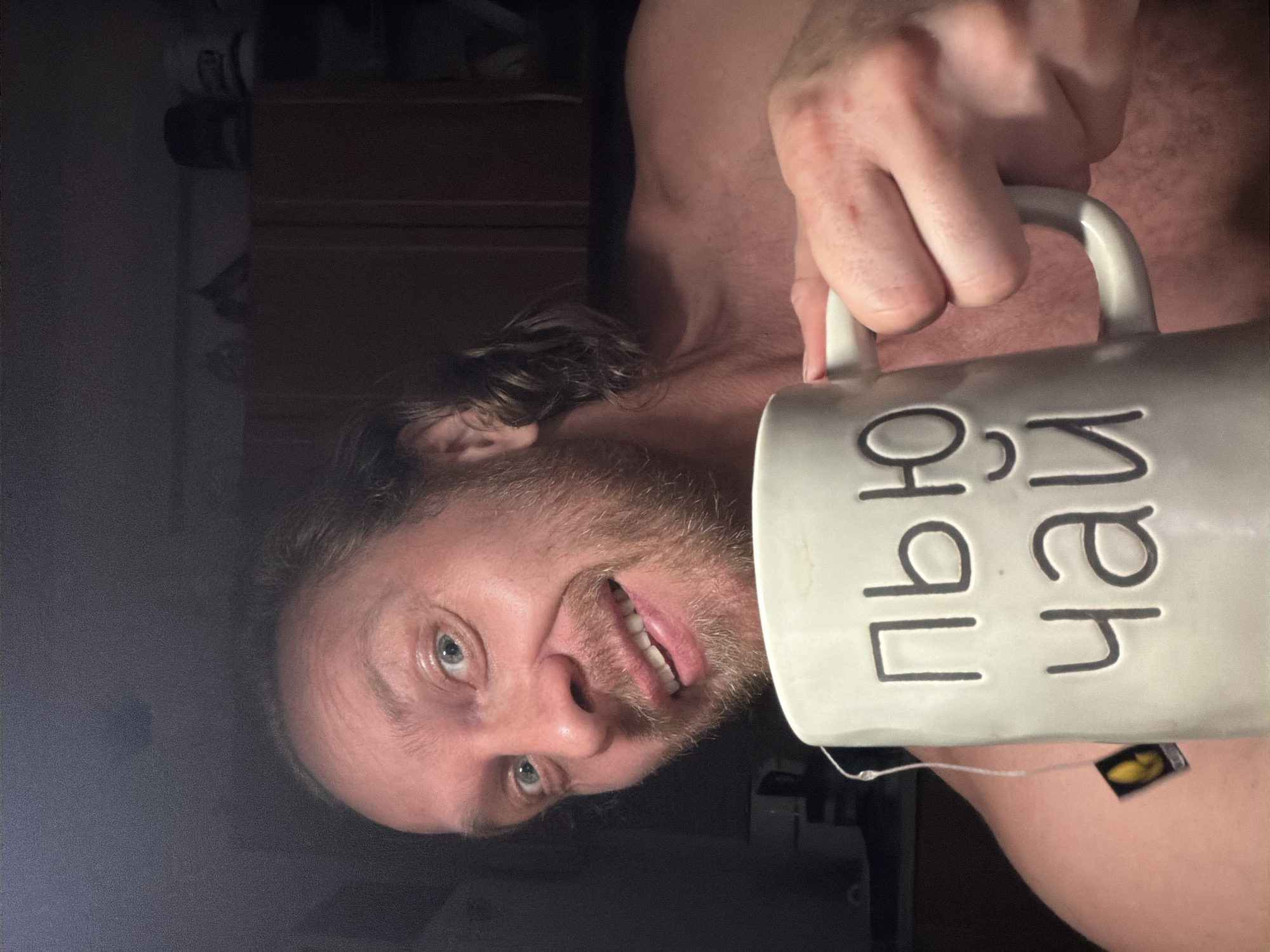 A man with light skin, a beard, and light brown hair, is holding a white mug with Cyrillic text that translates to "I drink tea." The mug has a tea bag hanging from its side. The man is shirtless and appears to be indoors in what looks like a kitchen with cabinets in the background. The scene is lit with artificial light and no specific location in Saint Petersburg is identifiable.