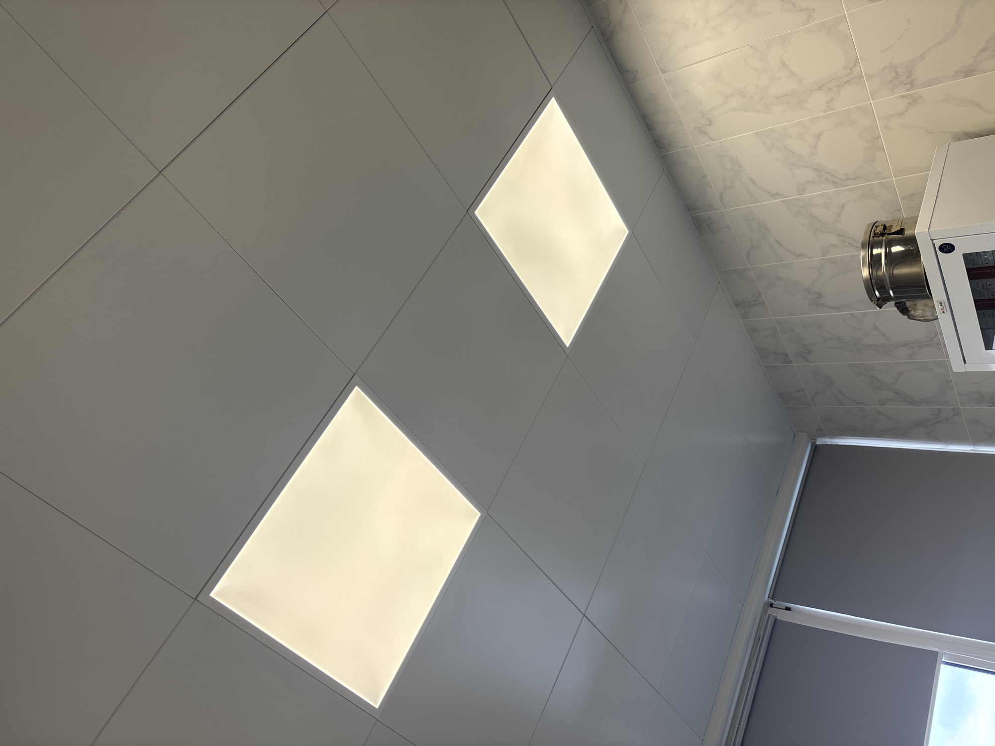 The image shows an upward angled view of a room's ceiling and the upper portion of one wall. The ceiling is composed of a grid of square gray tiles, with two square recessed lights illuminating the scene. The floor near the wall is covered with white marble tiles. The visible wall includes a gray roller shade covering a window. A metallic object with ductwork and a white appliance are partially visible in the top right corner. The image location appears to be indoors. The location is unknown.