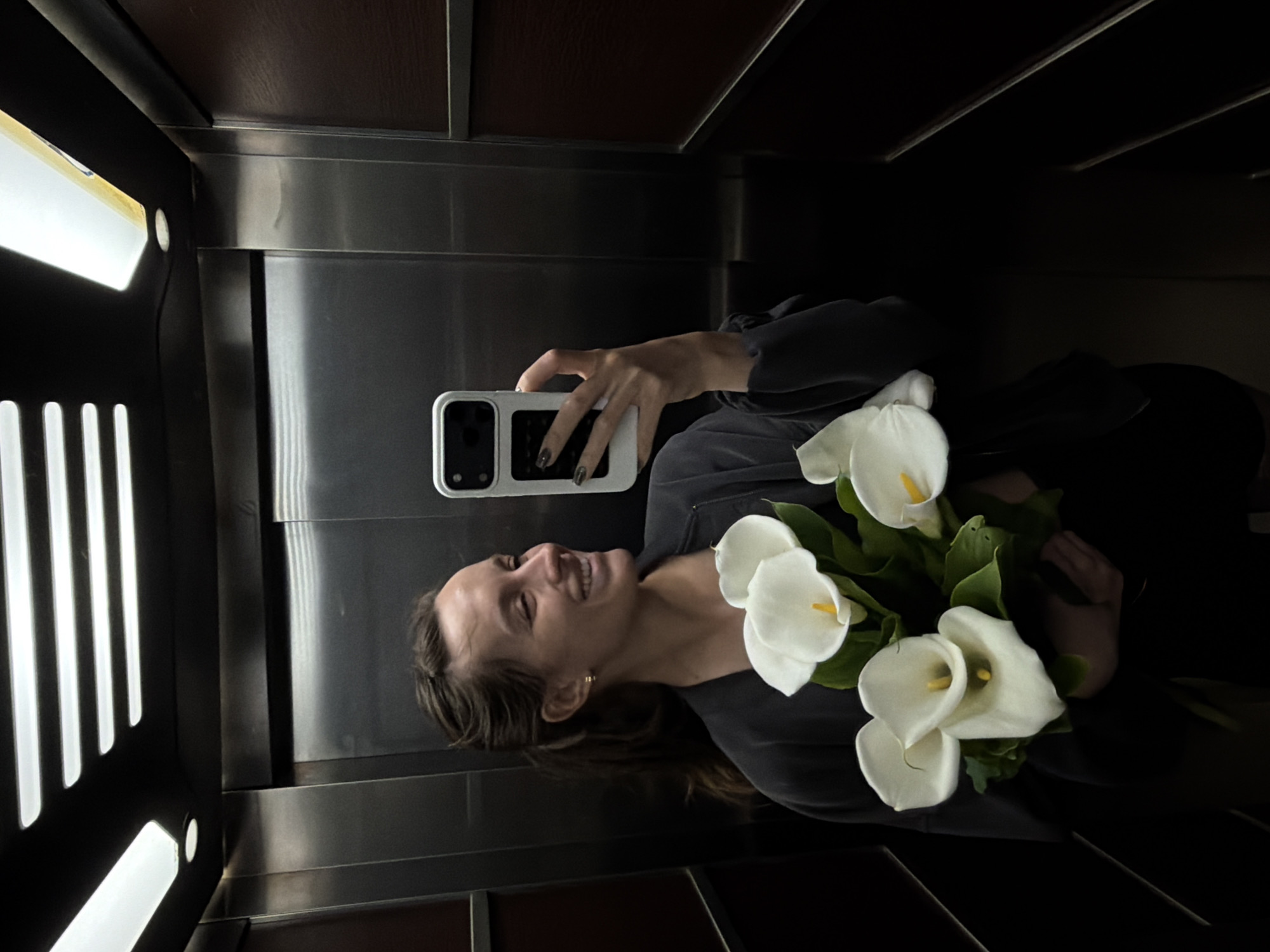 A woman with long brown hair smiles broadly while taking a mirror selfie inside an elevator in Budva, Montenegro. She holds a bouquet of white calla lilies with prominent yellow spadices and green leaves in her left arm, and a light-colored smartphone with a dark camera module in her right hand, positioned to capture her reflection. Her fingernails are painted a dark color, and a small gold hoop earring is visible on her right ear. She is wearing a dark, long-sleeved top. The elevator's interior features metallic paneling on the walls, with darker, possibly wooden, panels at the top and bottom. Bright artificial light emanates from a vertical fixture on the left side of the elevator, reflecting off the metallic surfaces. No discernible text is visible. The time of day is not evident due to the indoor, artificial lighting.
