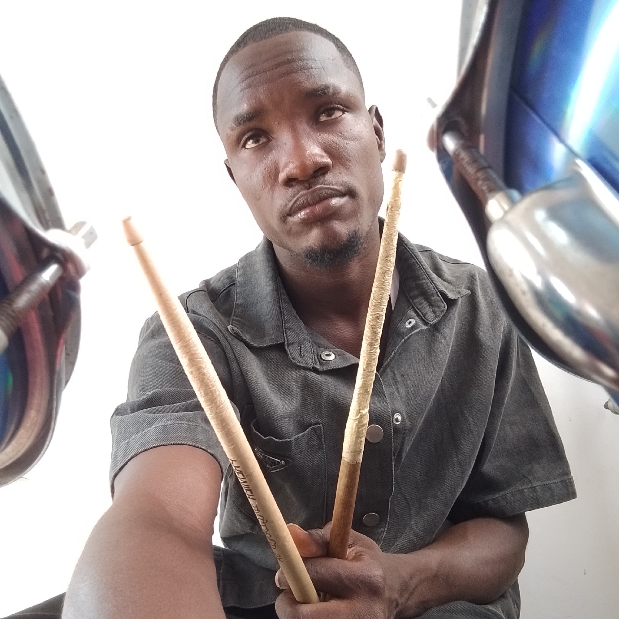 The image, taken in Iranla, Nigeria, shows a close-up of a Black man holding two drumsticks. He is wearing a dark grey denim shirt with the collar unbuttoned and metal snaps down the front. The man has short hair and a goatee. His gaze is directed upwards. On the left and right edges of the frame, parts of what appear to be cymbals or other metallic drum kit components are visible, reflecting a blue color and the bright white background. The man's hands are holding the drumsticks, with one stick showing visible lettering. The overall impression is that of a musician preparing to play.