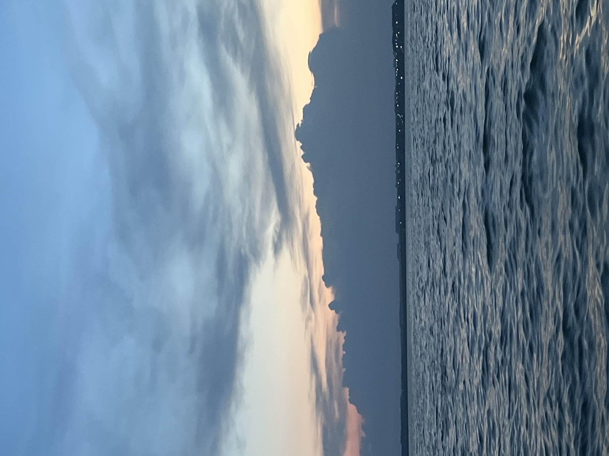 A serene sunset scene unfolds over a body of water, possibly the Ob River, as viewed from Novopichugovo, Russia. The sky is a canvas of blues and grays, with soft hues of pink and orange peeking through at the horizon. A thick, dark cloud formation dominates the mid-ground, casting a dramatic silhouette against the fading light. Below this, a distant shoreline is visible, dotted with faint, twinkling lights that suggest a settlement or town. The foreground is a expanse of calm water, its surface rippling gently, reflecting the muted colors of the sky and clouds. No people or specific actions are depicted in the image. The overall atmosphere is one of tranquility and natural beauty.