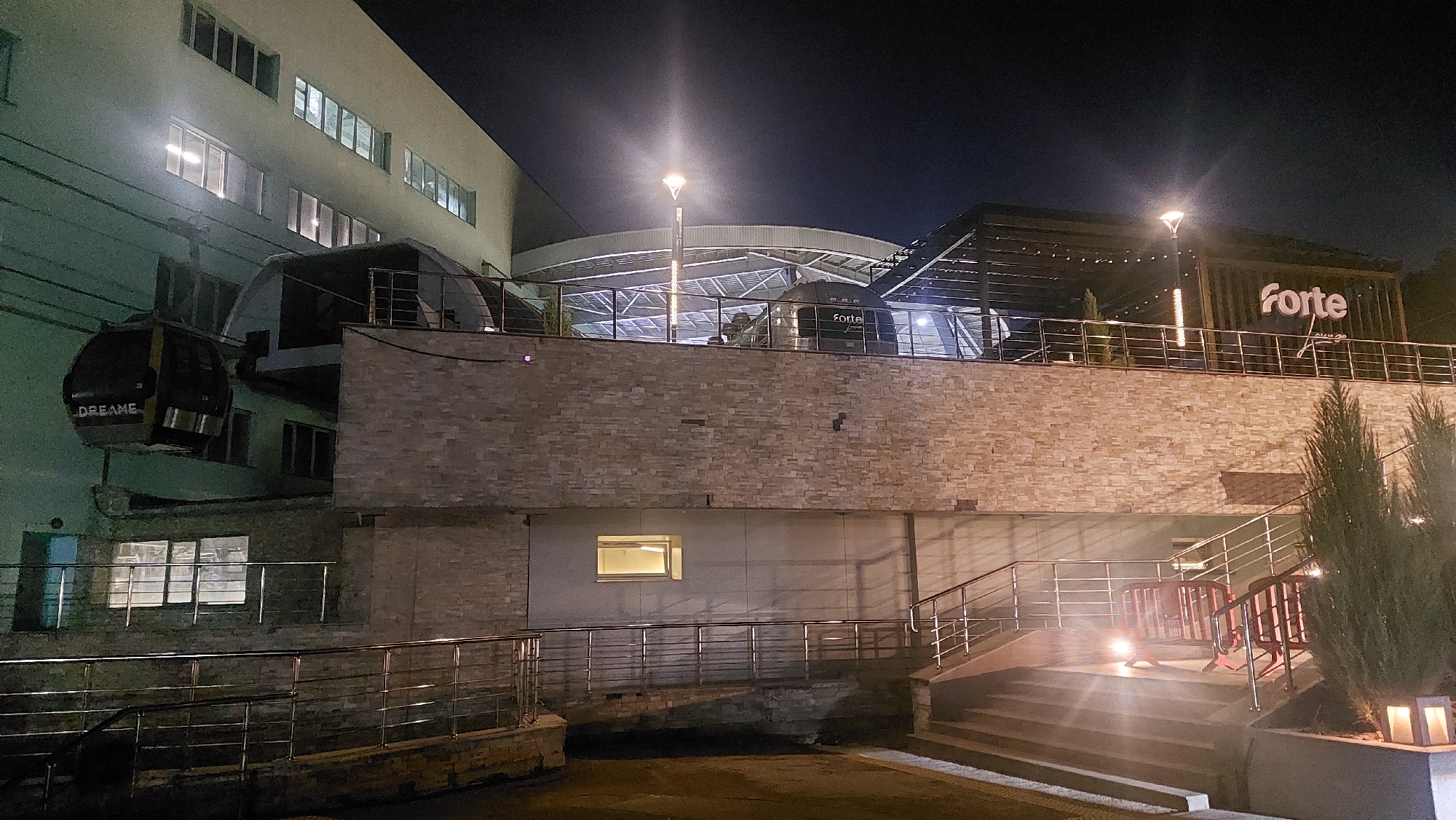The image depicts a night scene at the "Forte Lounge" in Almaty, Kazakhstan. A cable car, labeled "DREAME," is visible on the left, suspended by wires. The lounge itself is a multi-story building with a contemporary design, featuring a prominent "forte" sign illuminated at night. The building has a brick facade on the lower levels and glass-enclosed upper sections. Several streetlights are visible, casting a glow on the surroundings. A set of stairs leads up to the entrance of the lounge, with some red barricades placed near the bottom of the stairs. The overall atmosphere is that of a modern establishment in an urban setting at night.