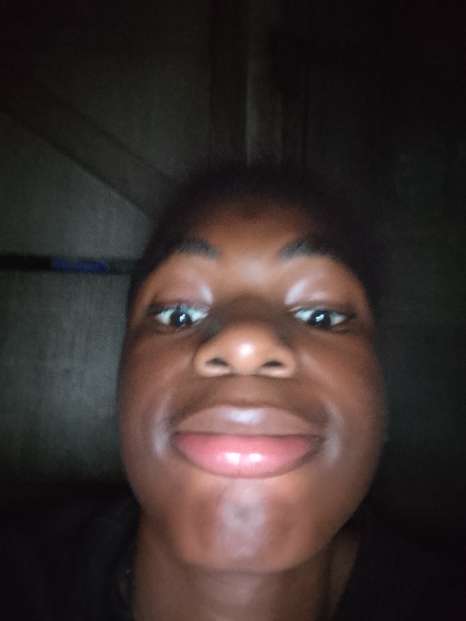 A close-up, low-angle portrait features a person with dark skin looking directly into the camera. The subject's face dominates the frame, with wide, dark eyes reflecting a faint light source. A broad nose is visible, and the lips are full, slightly parted, and have a pinkish hue, suggesting a relaxed or subtly smirking expression. The lighting is dim, casting soft shadows on the face and creating a low-key atmosphere.
The background is very dark and mostly indistinct, with faint vertical lines or textures hinting at a wall or door behind the subject. A blurry, elongated dark blue object, possibly a pen or similar item, is faintly visible horizontally in the upper left background behind the head. The person's dark hair blends into the dark surroundings. The image appears to be a selfie taken in a dimly lit indoor setting. No text is visible.