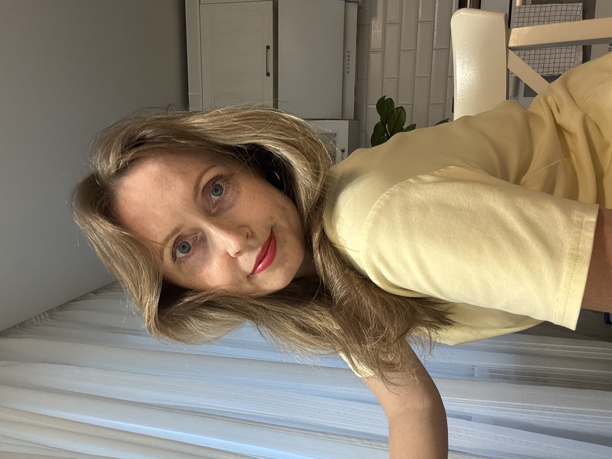 A woman with long blonde hair and red lipstick is in an indoor setting. She is wearing a pale yellow shirt. In the background, there is a white cabinet and white tiled wall with a potted plant. A white chair is partially visible. The location is the city of Chaikovsky, Russia.