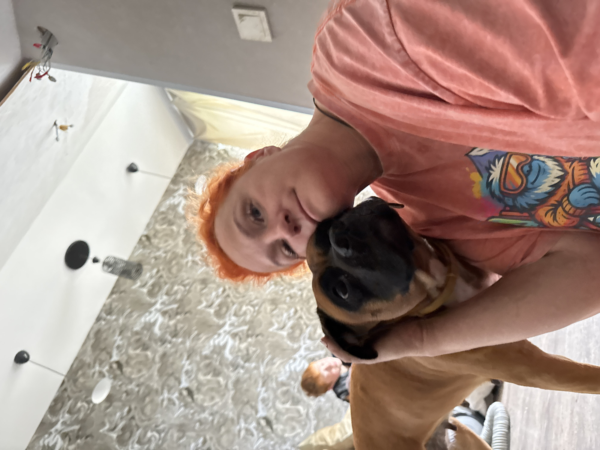 A woman with bright orange hair is taking a selfie with her brown boxer dog. The dog is leaning its head into the woman's shoulder, and the woman has her arm around the dog. In the background, a young boy with red hair is visible. The scene appears to be indoors, possibly a living room, with textured wallpaper and ceiling lights. There are also some decorative items on the ceiling, including what looks like a miniature airplane. The woman is wearing a coral-colored t-shirt with a graphic print on the front. The overall impression is one of a warm and affectionate moment between the woman, her dog, and her child. The image is taken from a low angle, looking up towards the ceiling.