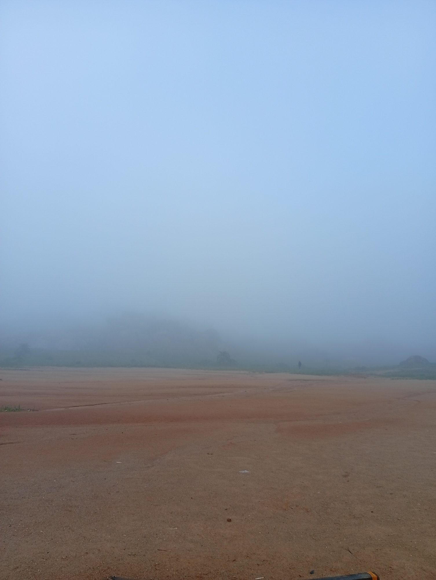 The image depicts a wide, open, sandy area with a person standing in the distance, partially obscured by fog. The ground is a reddish-brown sand, with some sparse vegetation visible in the background. The sky is a pale blue, merging into the thick fog that blankets the landscape. The fog reduces visibility significantly, obscuring most of the background details. The person appears to be walking or standing alone in the vast, misty expanse. The scene has a serene, somewhat desolate atmosphere due to the fog and the solitary figure. The location is identified as the city of Jos, Nigeria.