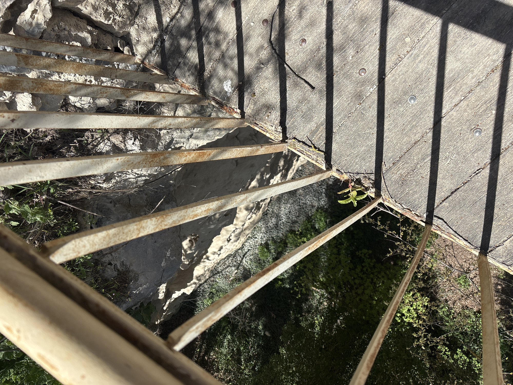 View from above of rusted metal railing on concrete and stone structure. Below the structure is exposed stone and vegetation. Light and shadow are visible throughout the scene. Location: Unknown, Unknown.