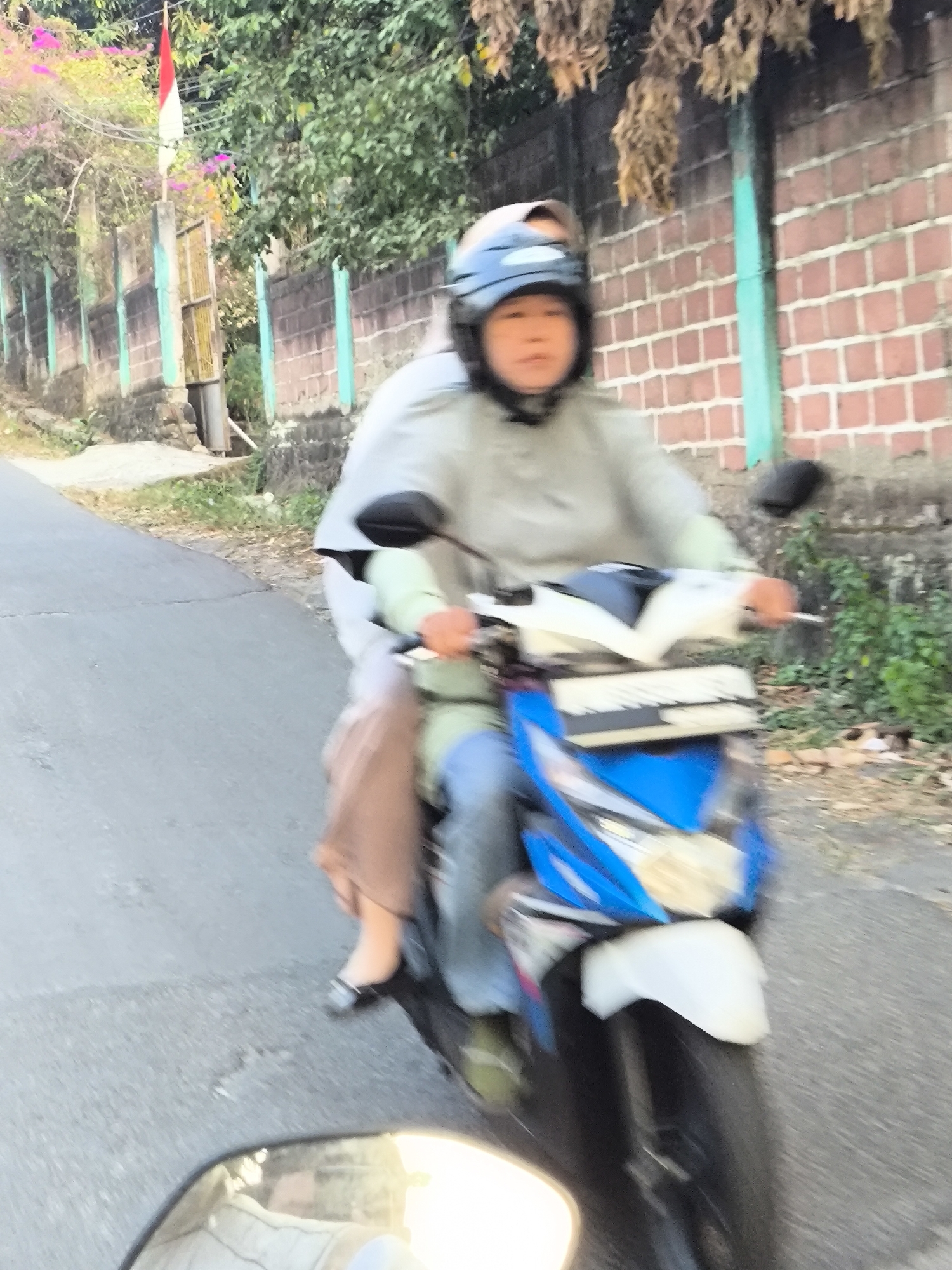 In the city of Cilegon, Indonesia, a person wearing a helmet rides a motorcycle on a paved road. The motorcycle is blue and white with a black fender. The rider is wearing a light-colored jacket and blue pants. A second person is riding as a passenger, wearing a brown skirt and a white shirt. The road is lined with a brick wall and green vegetation. A red and white flag is visible in the background. The scene appears to be captured in daylight.
