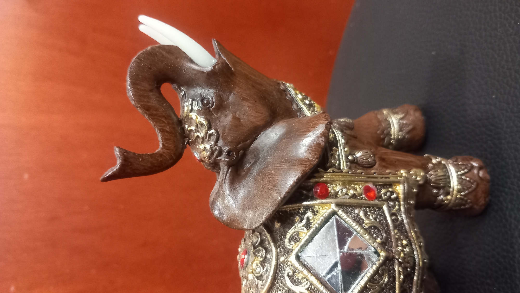 A close-up, medium-shot photograph shows a decorative elephant figurine against an out-of-focus, reddish-orange background. The figurine is positioned on a dark, textured surface, likely a table or stool. The elephant is brown, with ornate gold and silver detailing, including a saddle-like cover adorned with mirrored squares and red jewel-like accents. Its trunk is raised, and it has white tusks. The lighting is consistent, highlighting the detail and texture of the figurine. There is no indication of the city of Kos, Greece in the image.