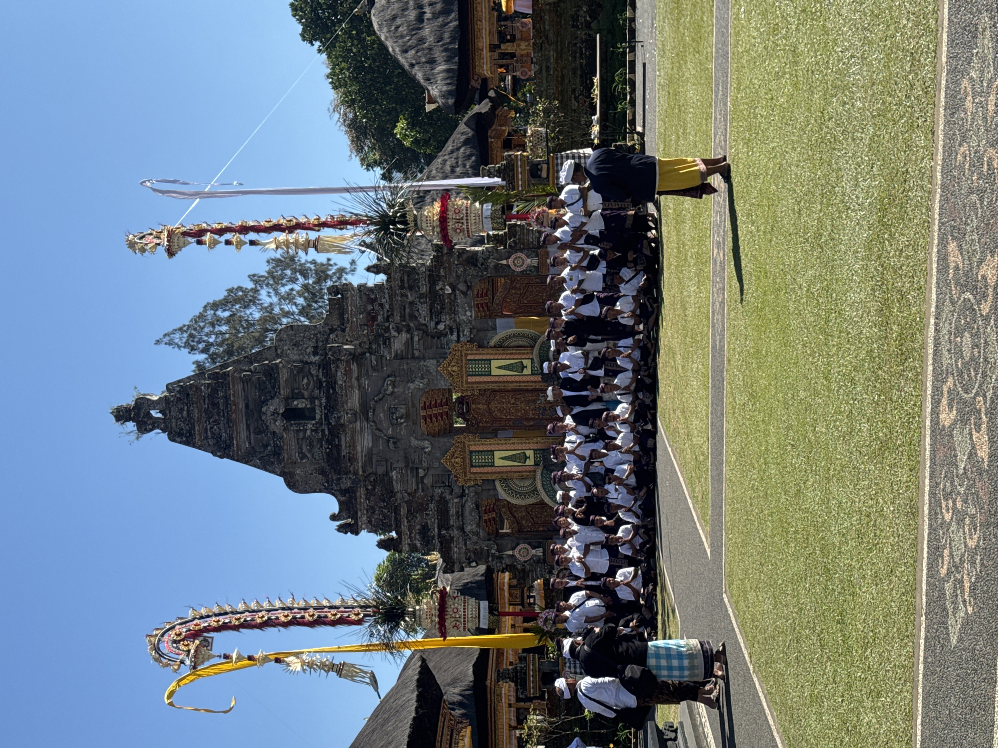 This image captures a vibrant outdoor Balinese ceremony taking place on a clear, sunny day. In the foreground, a large group of people, predominantly men, are seated in neat rows on a green lawn, all facing towards a traditional Balinese temple (Pura) in the background. They are dressed in ceremonial attire, including white shirts, dark sarongs (kain), and white head coverings (udeng).
The temple itself is an ornate stone structure featuring intricate carvings and multiple tiered roofs (meru). Flanking the seated congregation and near the temple are several tall, elaborate ceremonial poles known as penjor. These are richly decorated with long white and red fabric streamers, yellow ribbons, and traditional offerings.
Two individuals stand prominently; one on the far right, wearing a yellow sarong and dark top, appears to be observing or participating in a leadership role. Another person in traditional attire is partially visible on the far left. The bright sunlight casts strong shadows, indicating it is likely midday or early afternoon. The background features a clear blue sky and lush green trees. Paved walkways with subtle patterns are visible on the right side of the lawn. The overall atmosphere suggests a solemn and culturally significant religious event in Baturiti, Indonesia. No discernible text is visible.