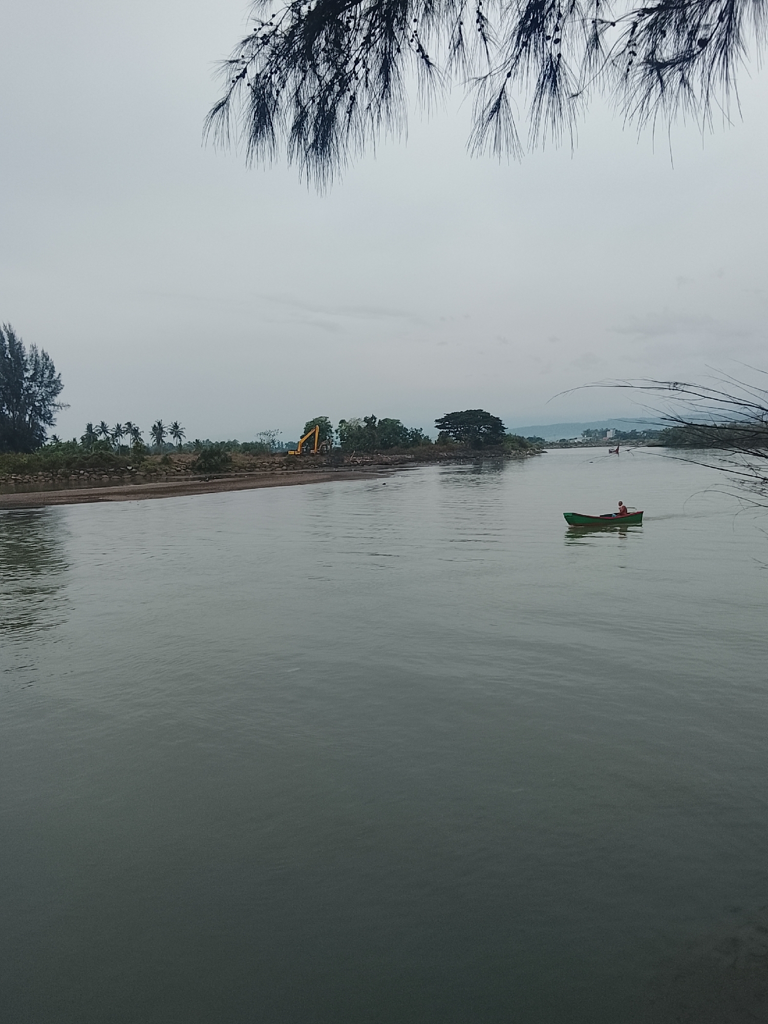 This is a horizontal, medium-distance, eye-level photograph of a river in Reudeuep, Indonesia. The sky is overcast and pale gray, with dark, needle-like branches of a tree hanging from the top of the frame. The river occupies the majority of the lower half of the image and is a muted green-gray color with subtle ripples. On the left bank of the river, there is a sandy shore, followed by a row of palm trees and other vegetation. An excavator is visible on the shore in the distance. Further down the river, on the right bank, a small green boat with a person wearing an orange shirt is moving away from the camera. The river narrows in the distance, leading to a more developed area with buildings visible. The overall mood is subdued and natural.