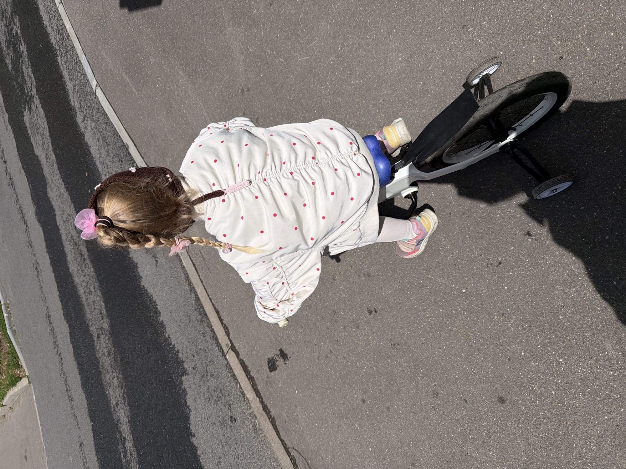 A young girl with blonde hair in braids, adorned with pink bows and a brown knitted hat, rides a white balance bike with training wheels. She is wearing a white jacket with red polka dots and pink and yellow sneakers. The girl is on an asphalt surface, likely a sidewalk or bike path, with a curb and grass visible to the left. The scene is set outdoors on a sunny day, casting a shadow of the girl and the bike onto the ground. The image appears to have been taken in Moscow, Russia, based on context.