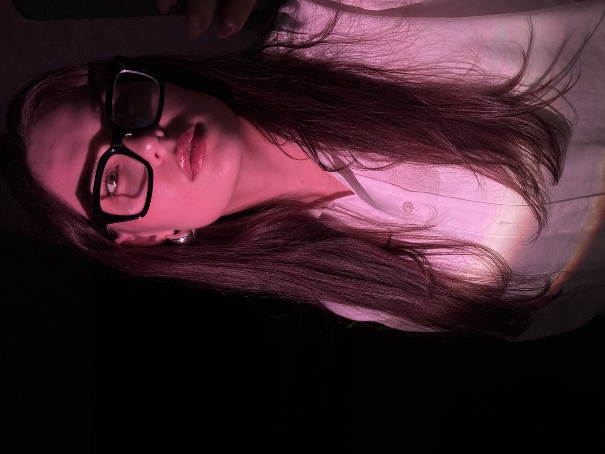 The image shows a close-up of a young woman wearing glasses and a white button-down shirt. She has long, dark hair that is illuminated by a pink light, casting shadows and creating a dramatic effect. The background is mostly dark, with a hint of light on the right side of the frame. The woman is looking towards the camera with her lips slightly parted. There is no indication of the location being Chaikovsky, Russia, or any other specific city.