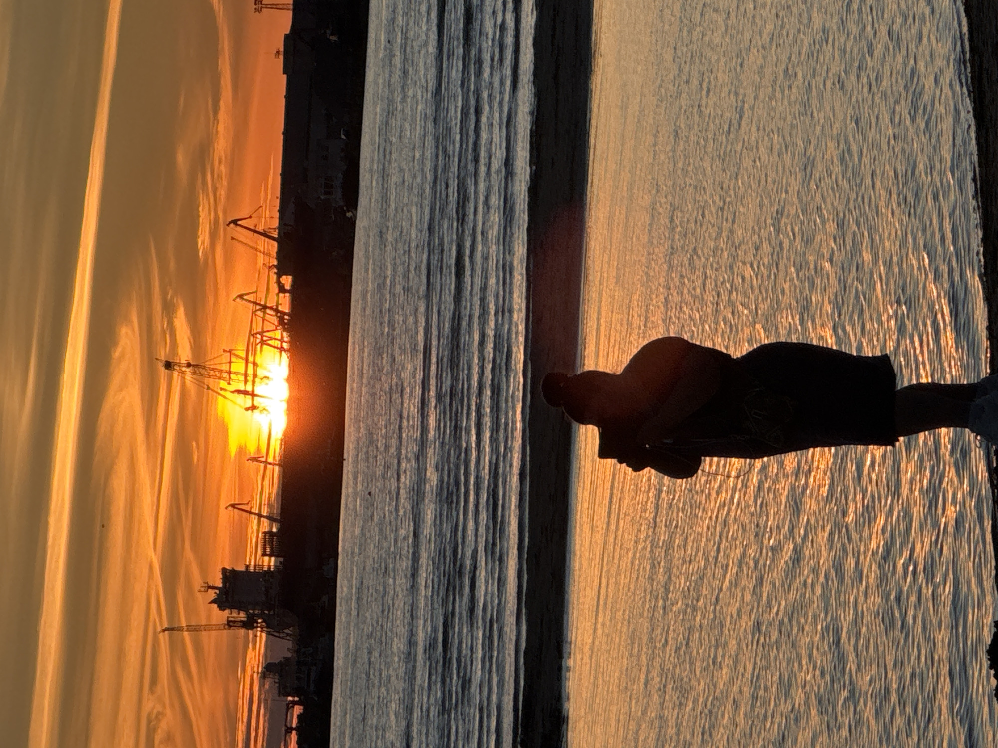 A silhouette of a person standing in shallow water at the edge of a beach holds a phone up to take a picture of a sunset over a harbor. The sun is setting behind a line of industrial cranes and ships, casting a vibrant orange and yellow glow across the sky and reflecting on the water. The person is facing away from the camera and their back is to the viewer. The image appears to be taken at sunset from a beach in Tallinn, Estonia.
