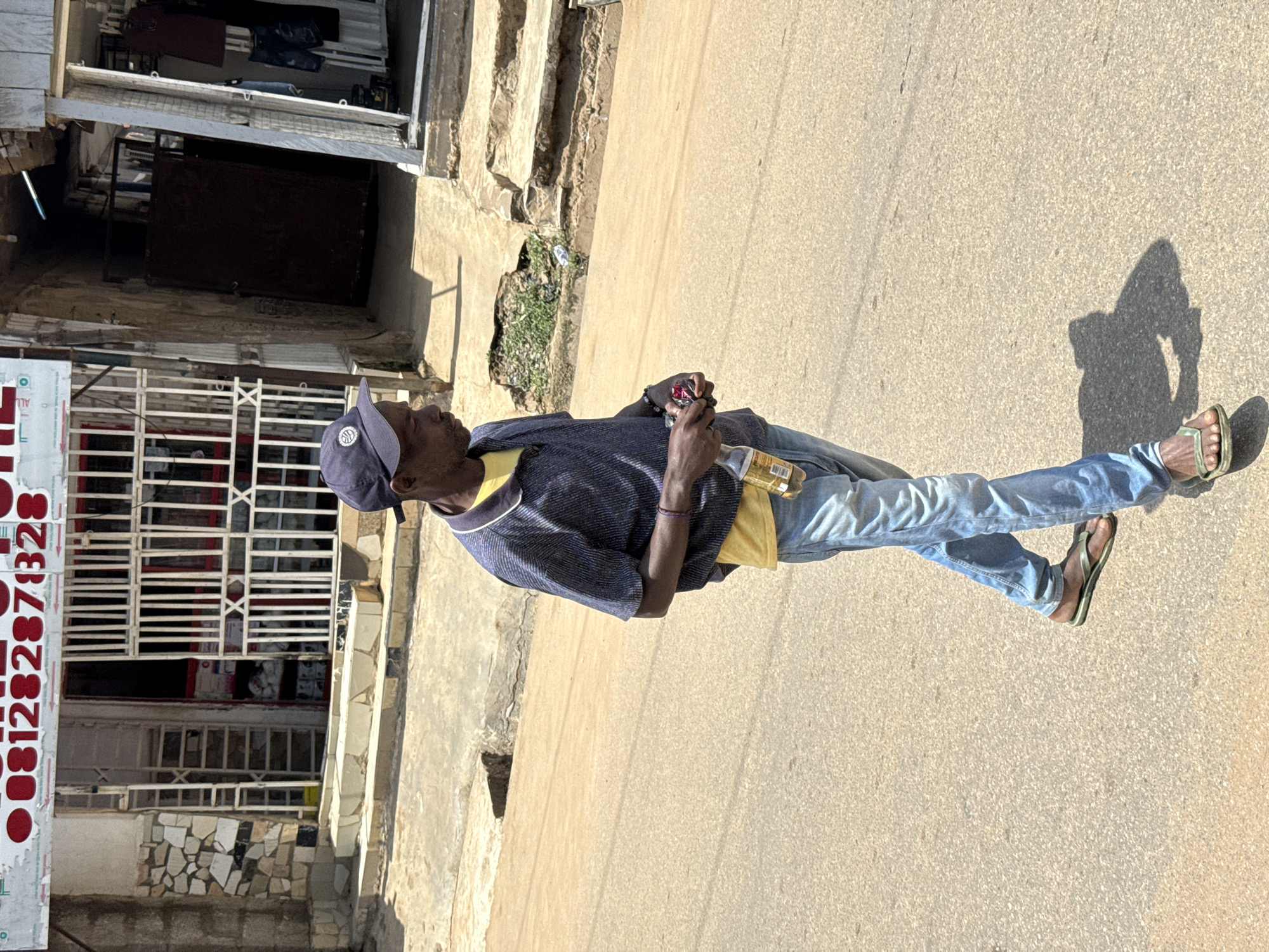 A man, appearing to be middle-aged or older, stands on a dusty street in what appears to be a commercial area. He is wearing a dark blue shirt with a yellow collar, light blue jeans, a navy baseball cap, and brown flip-flops. He is holding a small, dark object in his right hand and a bottle in his left. His facial expression is neutral, and the overall impression is of a person standing idly on the street.
The scene is set against a backdrop of buildings with storefronts. One storefront on the left has a white metal grille and a sign with red lettering that appears to be a phone number: "081282871328". The buildings and street suggest a sunny day, possibly late morning or early afternoon, given the strong shadows cast on the ground. The environment appears somewhat worn, with visible cracks in the pavement and a patch of overgrown grass near one of the buildings. The general atmosphere is that of a typical street scene in a Nigerian city like Abuja, with the presence of shops and the unpaved or rough asphalt of the road. There are no other people clearly visible in the immediate vicinity, though there might be activity within the shops.